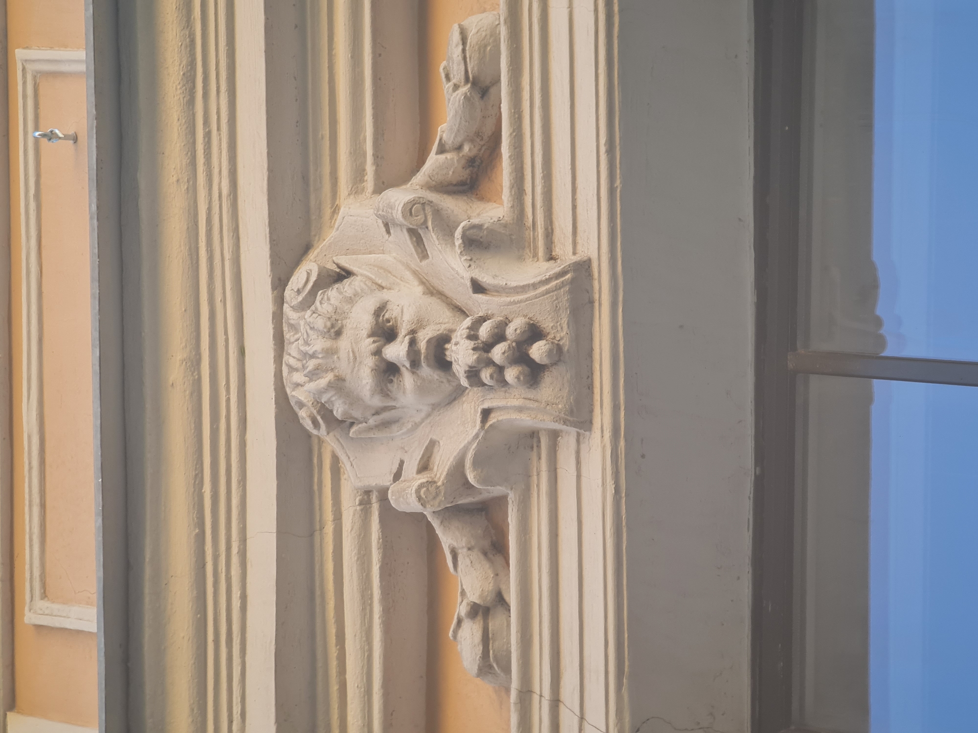 A stone relief of a grotesque face is mounted above a window in Saint Petersburg, Russia. The face, with its bulging eyes and open mouth, appears to be spitting out a cluster of grapes. The relief is surrounded by ornate architectural elements, including carved leaves and scrolls. The building facade is painted a pale yellow, and the relief is a light, creamy color. The window below reflects a clear blue sky. The overall scene suggests an old, possibly historical building, typical of the architecture found in the city's historic center.