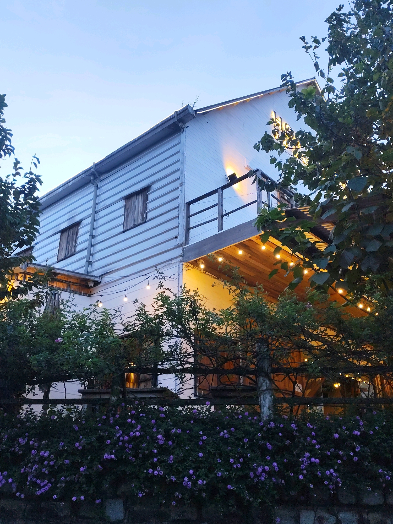 The image shows a two-story building with white horizontal siding, possibly made of wood or vinyl, set against a twilight sky. The building has two small, wooden-shuttered windows on the upper level. A balcony with a wooden railing extends from the second story, and a wooden deck is visible underneath the balcony, illuminated by string lights. More string lights are draped along the edge of the balcony railing. Green trees and bushes with small purple flowers partially obscure the lower part of the building and the fence in front of it. The overall scene suggests an outdoor seating area or a quaint establishment, possibly a cafe or restaurant, in Da Lat, Vietnam, given the style of architecture and the lush greenery common in the region. The lighting and the twilight sky indicate the time is early evening. No people are visible in the image.