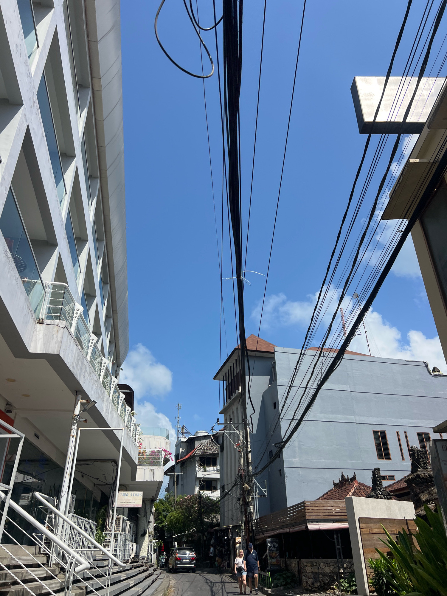 The image shows a street scene in Kuta, Indonesia, under a bright blue sky with scattered white clouds. The view is looking up at the sky, dominated by a tangled network of overhead electrical wires.
On the left, a modern building with a white facade and numerous windows is visible. The windows are framed by diagonal concrete elements, giving the building a distinctive geometric pattern. A white railing lines a balcony or walkway on the lower level of this building.
To the right of the modern building, a narrow street leads into the distance. A light blue building with dark-tinted rectangular windows stands prominently on the right side of the street. Below this building, a lower structure with a wooden railing and a thatched roof suggests a restaurant or shop. Traditional Balinese architectural elements, such as stone carvings and tiled roofs, are visible on some of the buildings.
In the street, a silver car is parked, and two people are walking away from the camera. The street is lined with shops and businesses, indicated by signs and storefronts. The presence of numerous electrical wires strung across the street is a common feature of urban environments in this region. Green foliage is also visible, adding to the tropical feel of the scene. The overall impression is of a busy commercial street in a tropical city.