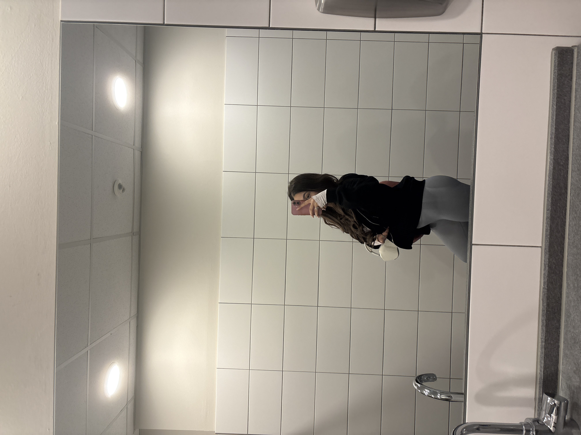 A woman wearing grey leggings and a black hooded jacket stands in a tiled bathroom, holding a pink cell phone and taking a selfie in the mirror. The bathroom appears to be in a commercial or public building. The ceiling features recessed lighting and white tiled panels. There is a sink with a faucet and a towel dispenser on the wall. The city is not discernable from the image.