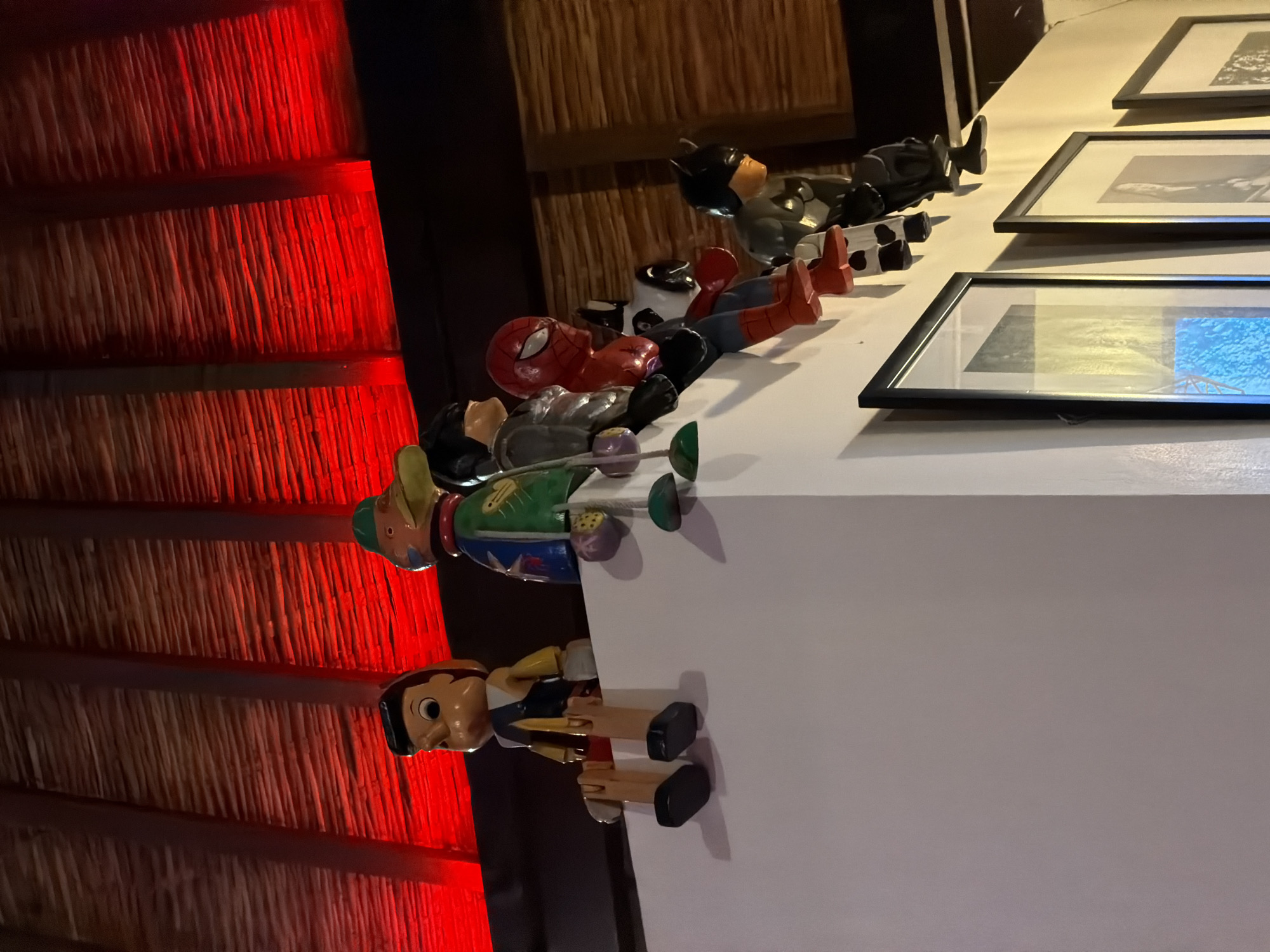 A still life photo shows a white ledge, likely a shelf, hosting a collection of character figurines and framed pictures. Starting from the front, a Pinocchio doll, wooden with movable limbs, stands on the ledge. Behind it is a wooden doll resembling a bullfighter. Further back are a couple of superhero figurines - Spiderman and Batman. Next to the figurines are three framed images, the content of which is not very clear. The background of the scene is split between what looks like a rustic bamboo wall and a wall illuminated by red light. The location is inferred to be West Denpasar, Indonesia, although there are no location-specific elements visible in the image to confirm.