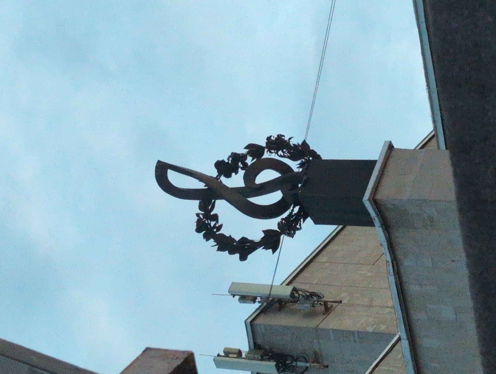 A low-angle shot depicts a sculptural element on a building in Izhevsk, Russia. The sculpture features a stylized rendering of the Udmurt symbol, a sign resembling an infinity symbol, surrounded by a wreath of metallic leaves. This decorative feature is mounted on a cantilevered architectural structure, possibly part of a larger building facade. Below the sculpture, on the building's wall, are several small, rectangular electronic components with connecting wires. The background is a pale blue sky. To the right and left of the sculpture are parts of the building structure.
