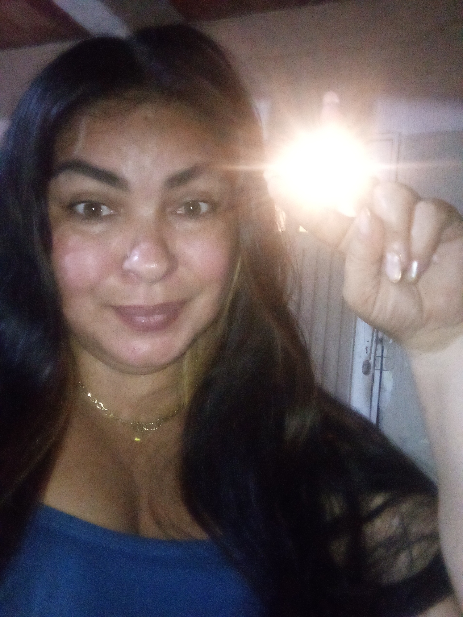 A female subject, appearing to be an adult, is positioned centrally, looking directly at the viewer with a slight smile. She has long, dark hair, brown eyes, and is wearing a blue garment and a thin gold-colored chain necklace with a small pendant. Her right hand is raised, holding a bright, self-generated light source, consistent with a smartphone flash, positioned to the right of her head. This light source creates significant glare and lens flare in the image, partially obscuring the background and her hand. The immediate environment appears to be an indoor setting, characterized by indistinct walls or surfaces in muted tones due to low ambient light and the flash. No specific identifying features of the city of Juan Griego, Venezuela, are discernible within the image.