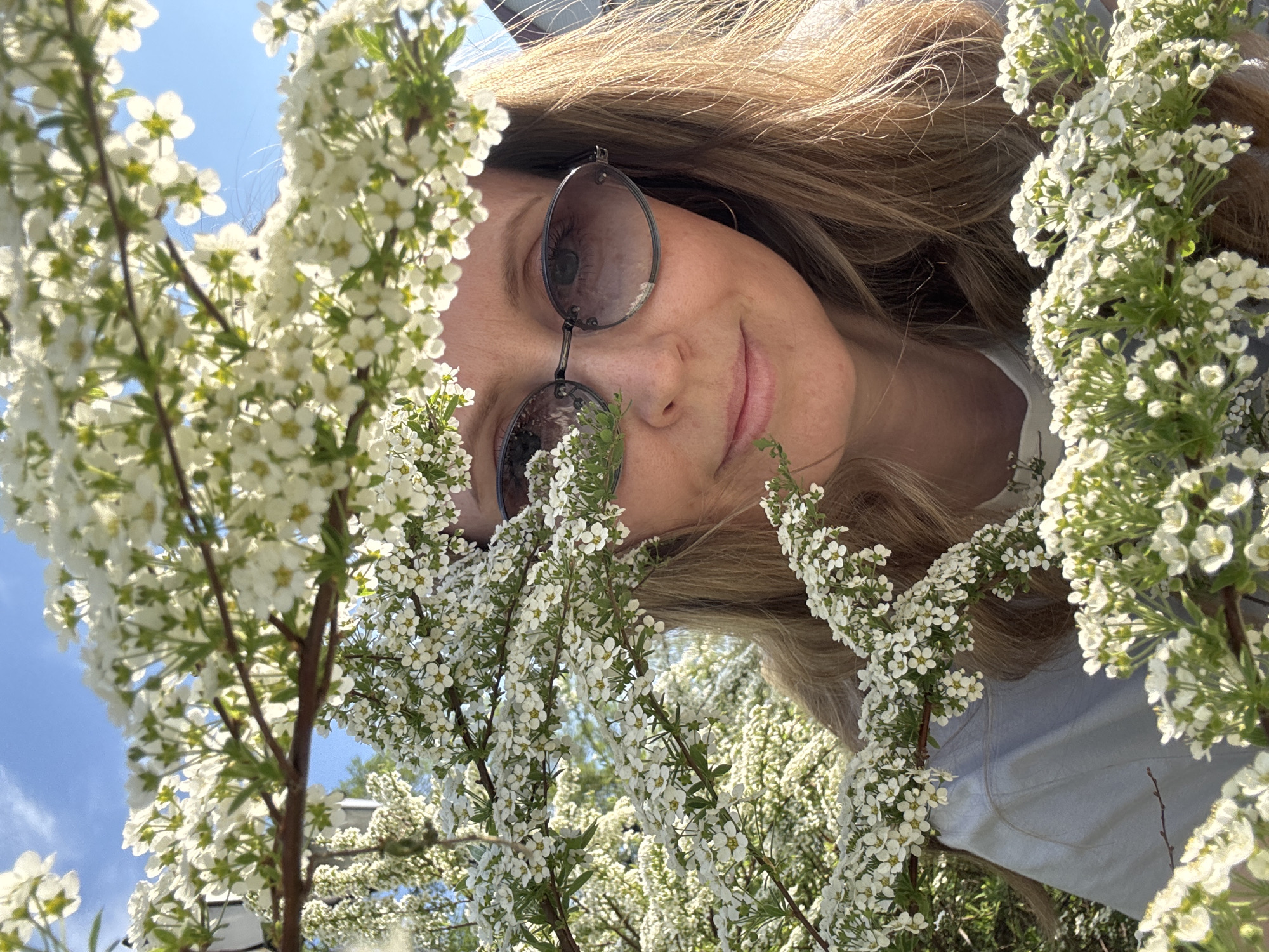 A woman with light brown hair and wearing sunglasses is positioned in a field of white flowers under a sunny sky. The photo is taken at an angle, with the flowers framing her face. The background hints at a bright blue sky with some greenery. Due to the lack of distinct landmarks or specific details, pinpointing the location within Votkinsk, Russia, is not possible.