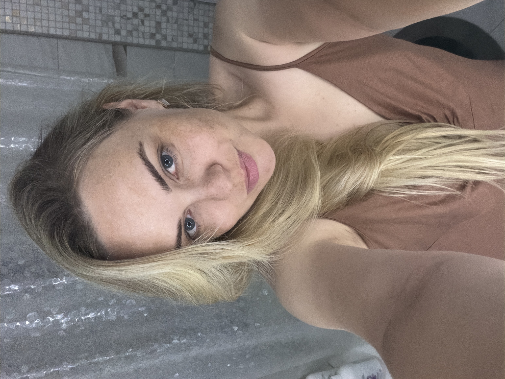 A woman with blonde hair and blue eyes poses for a selfie. She is wearing a brown tank top. The background appears to be a bathroom setting with tile visible. There is no information available to identify the location as the city of Votkinsk, Russia.