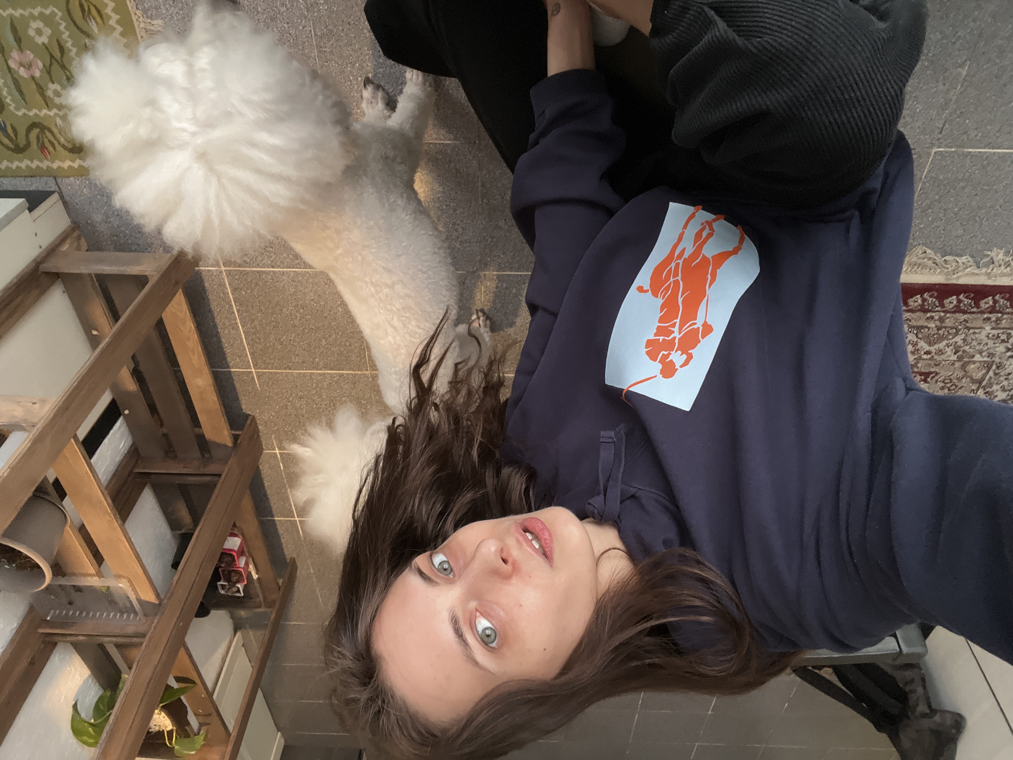 A woman with long brown hair and blue eyes lies on a gray tiled floor. She wears a navy blue hoodie with an orange Polo logo and black pants. A large white dog, possibly a poodle or similar breed, lies behind her. A wooden shelf unit with plants and other objects sits to her left. A rug is visible in the background. The location is indoors, possibly an apartment or home. There are no indications of Krasnoyarsk, Russia.