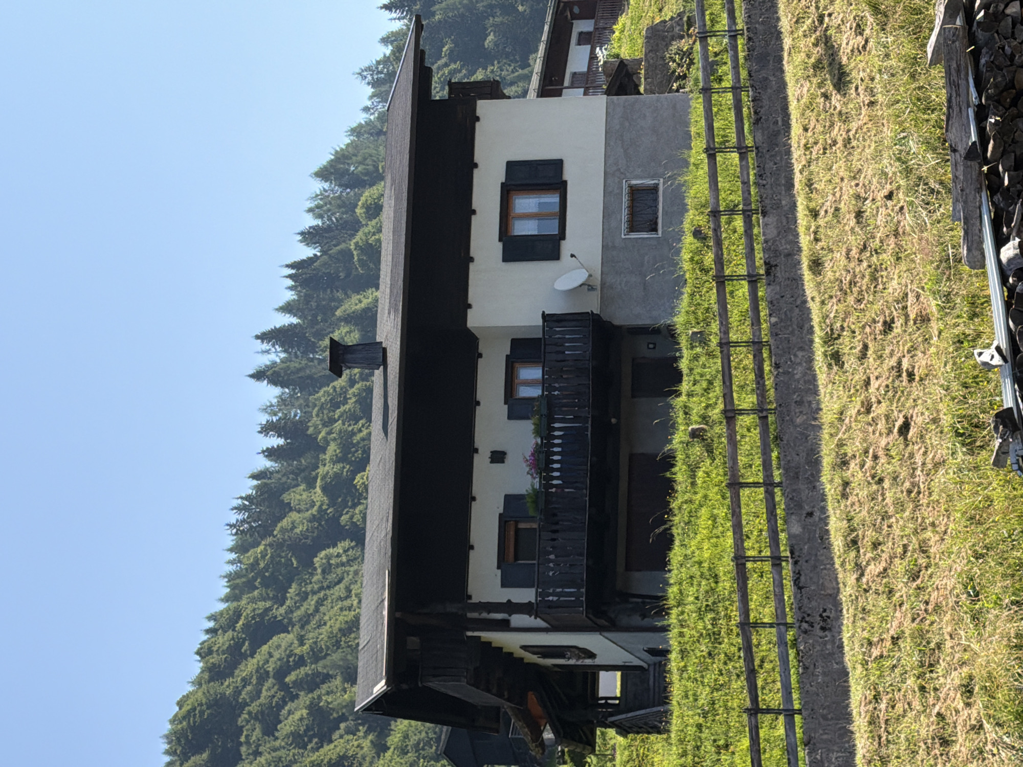 The image shows a multi-story building situated on a grassy hillside in Sauris / Zahre, Italy. The building features a dark-pitched roof with a chimney. The upper facade is white with dark-shuttered windows and a dark wooden balcony. The lower portion of the building is a light gray concrete. A wooden fence runs horizontally across the foreground, separating the building from a lower grassy slope. The background is dominated by a densely forested mountainside under a clear blue sky. No people or animals are visible in the scene.