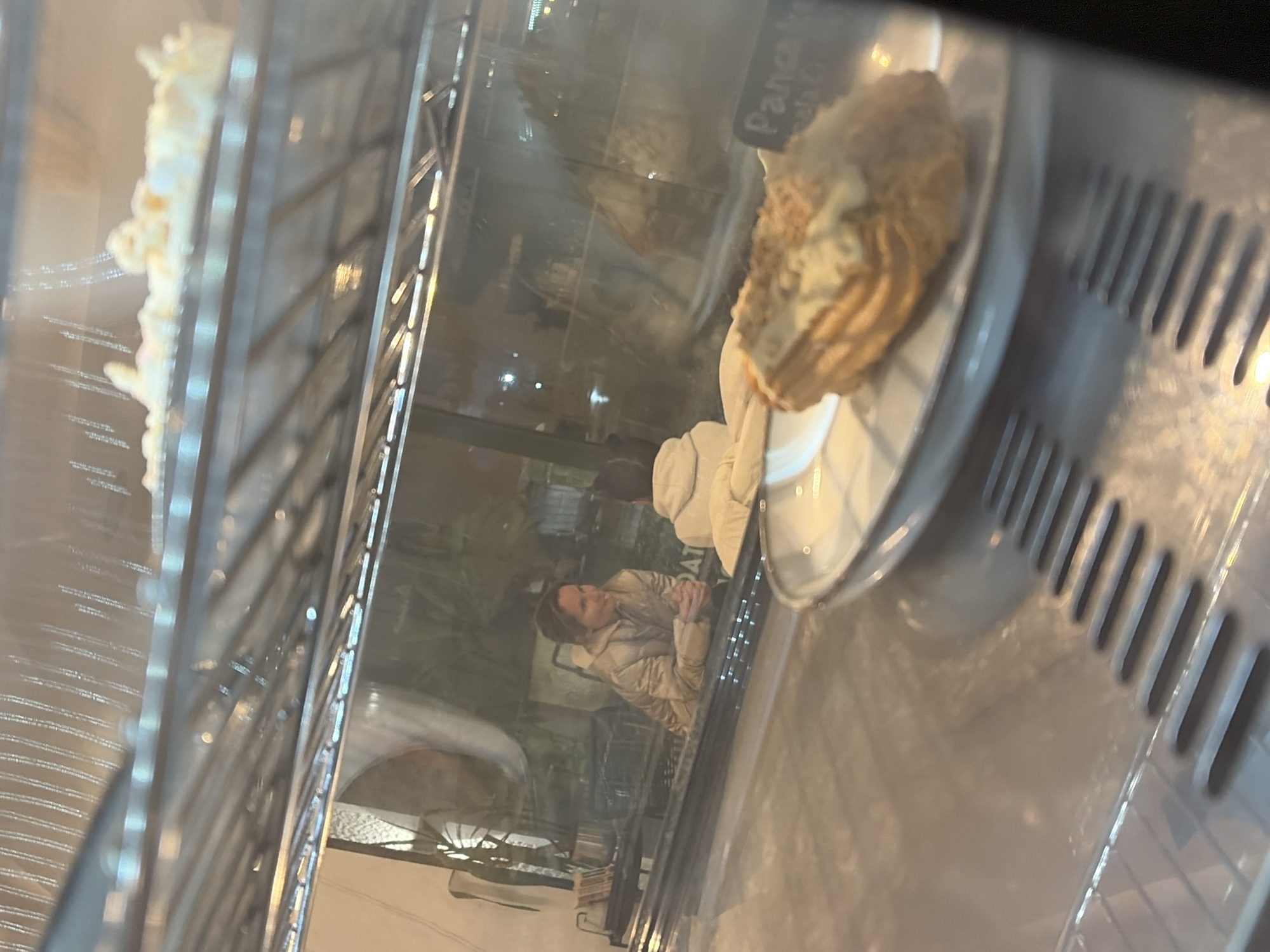 The image depicts an interior scene, viewed through the glass front of a chilled display case, likely in a cafe or bakery located in the city of Budva, Montenegro.
In the immediate foreground, a single oblong pastry, possibly an eclair or krempita, topped with light cream and finely crushed nuts, rests on a white plate on a lower metal shelf of the display case. A small, dark sign to the right of the pastry is partially visible, displaying text that includes "Pancake" or "Pahcka" at the top. Above this shelf, a reflective metal shelf holds a long, white dessert item, possibly meringue or marshmallow-based. The bottom interior of the display case features a ribbed metal surface.
Through the glass, reflecting or visible beyond the display, two individuals are seated at a table. A woman, positioned on the left, is seen from the chest up, wearing a light brown or beige puffer jacket. She has brown hair, appears to be smiling slightly, and has her hands clasped together on the table, indicating engagement in conversation. The second individual, to the right of the woman, is largely obscured from view, with only the back of their head and shoulders visible, wearing a light-colored hooded garment. Behind the seated individuals, a window or glass partition reveals a blurred outdoor or adjacent indoor area, with some illuminated greenery and distant lights, suggesting evening or night. The specific establishment or exact street location within Budva is not identifiable from the visual information provided.