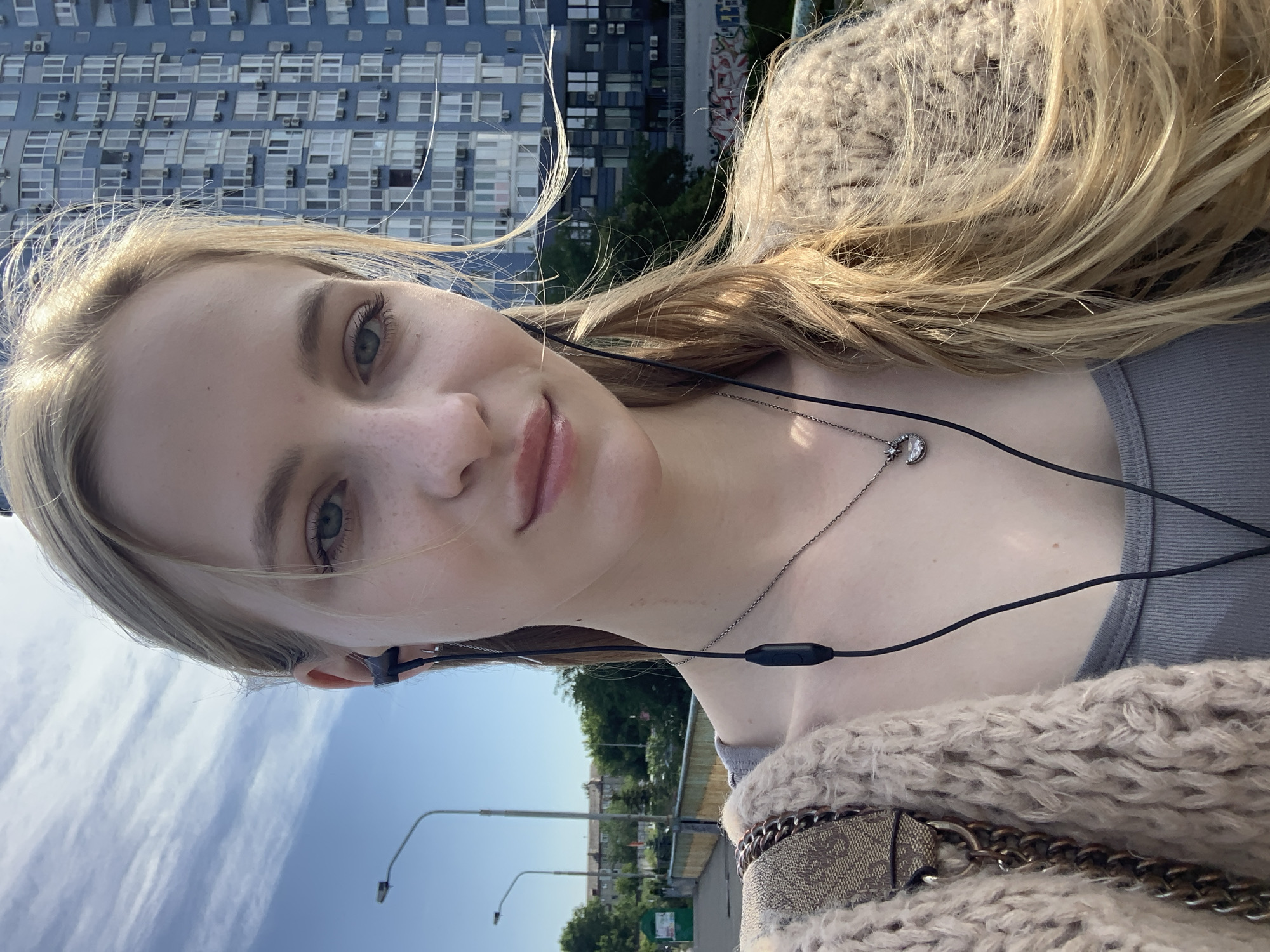 A young woman with blonde hair and blue eyes is taking a selfie outdoors on a sunny day. She is wearing a grey tank top, a beige knitted cardigan, and a necklace with a crescent moon pendant. She has black earbuds in her ears. In the background, there is a tall blue apartment building, a street light, trees, and a bridge. The overall environment suggests an urban setting, possibly in Kyiv, Ukraine, given the architectural style of the buildings and the general urban landscape. The woman appears to be in good health and is looking directly at the camera with a neutral expression.