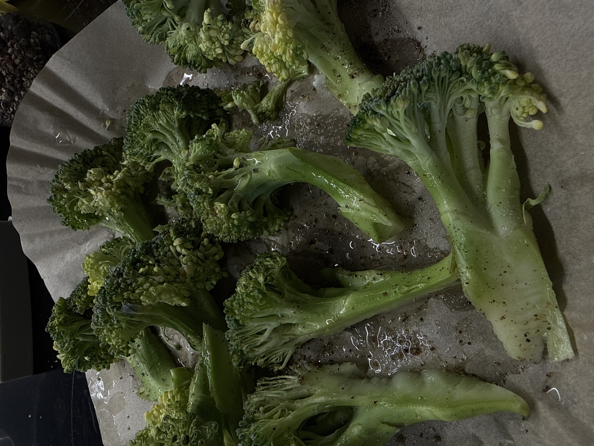 A close-up, high-angle shot captures numerous raw broccoli florets and stems arranged on a piece of light brown parchment paper. The broccoli pieces vary in size, with some larger florets showing distinct green crown structures and thicker stems, while smaller, more yellow-green pieces are also present.
The broccoli appears to be prepared for cooking, as evident by the droplets of liquid, likely water or cooking oil, glistening on the surface of the broccoli and the parchment paper. Small, dark specks, possibly ground black pepper or other seasoning, are scattered over the vegetable and the paper. The paper itself is wrinkled, suggesting it lines a cooking pan or tray, with some darker, possibly metal, surfaces visible around the edges of the paper in the background.
The lighting is somewhat dim, casting subtle shadows and highlighting the texture of the broccoli florets and the wet surfaces. No people are visible in the image, nor is there any discernible activity beyond the static arrangement of the broccoli. The setting appears to be an indoor kitchen environment. There is no visible text. The location context of Pervomaysky, Russia, cannot be confirmed from the visual information provided.