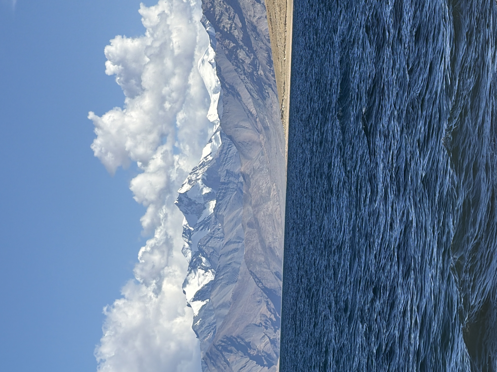 A vertical, medium shot captures a serene landscape scene. In the foreground, a vast expanse of deep blue water fills the bottom half of the frame. The surface of the water is rippled, indicating a gentle movement. Beyond the water, a dry, sandy shore stretches across the middle ground. This shore is dotted with a few small, light-colored objects that appear to be rocks or possibly grazing animals, though they are too indistinct to identify with certainty.
Dominating the background is a majestic mountain range. The lower slopes of the mountains are a muted brown and gray, showcasing their rugged texture. Higher up, these peaks are dramatically capped with pristine white snow, contrasting sharply with the darker rock. The mountains are partially obscured by a formation of fluffy white clouds against a bright blue sky. The clouds are scattered, with some appearing more dense and textured than others. The overall scene suggests a remote, natural environment, likely in a high-altitude region of India, given the presence of snow-capped mountains and the dry landscape.