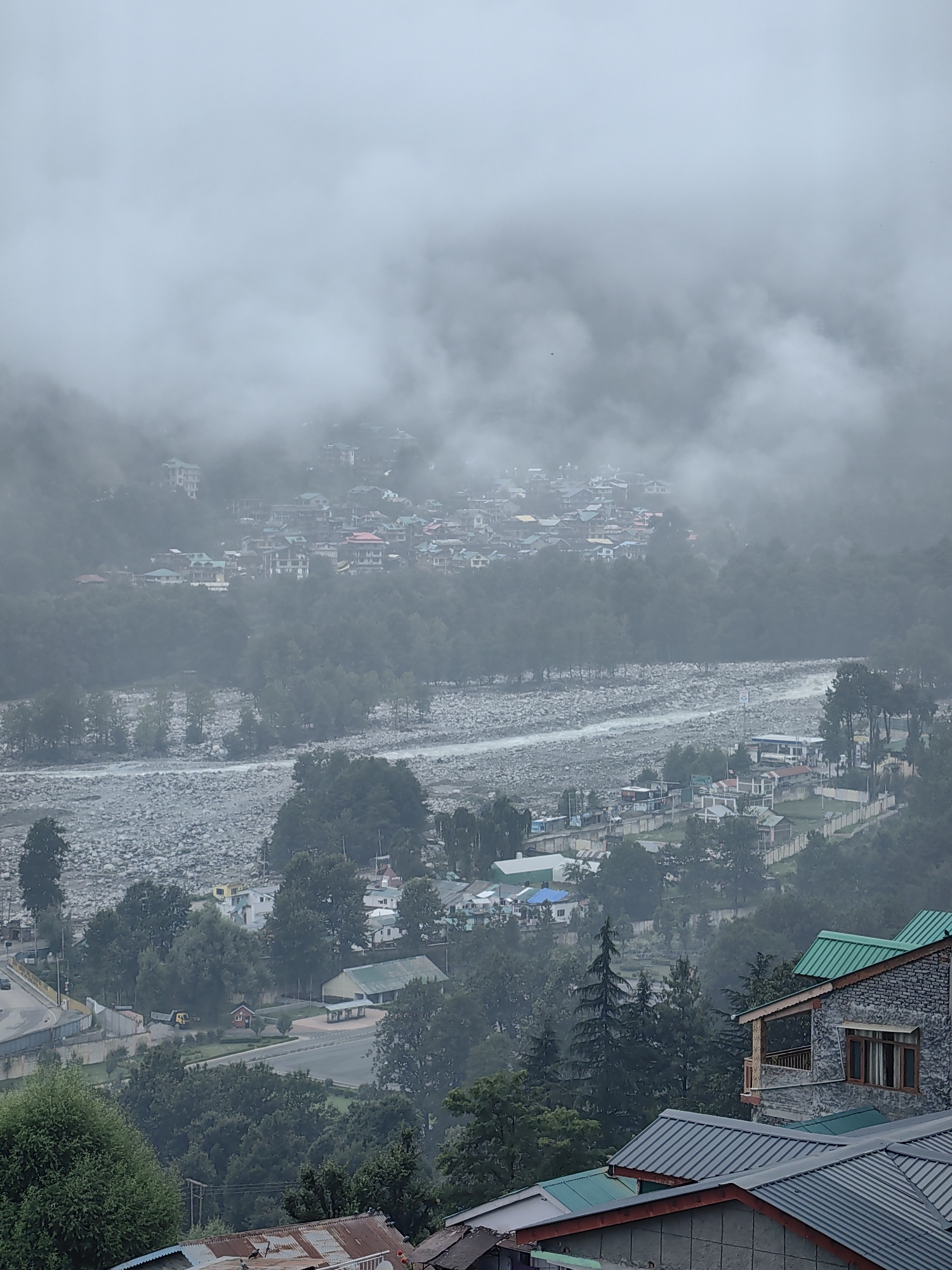 A misty, overcast sky hangs over a town nestled in a valley. In the foreground, two buildings with green metal roofs are visible. The building on the right has stone walls and a wooden balcony with railings. Beyond these buildings, a town spreads across the hillside, characterized by numerous houses with red, green, and blue roofs. A river with a rocky bed flows through the valley, bordered by trees. Further up the hillside, more buildings are visible, partially obscured by fog. The scene appears to be a view of Vashisht, India.