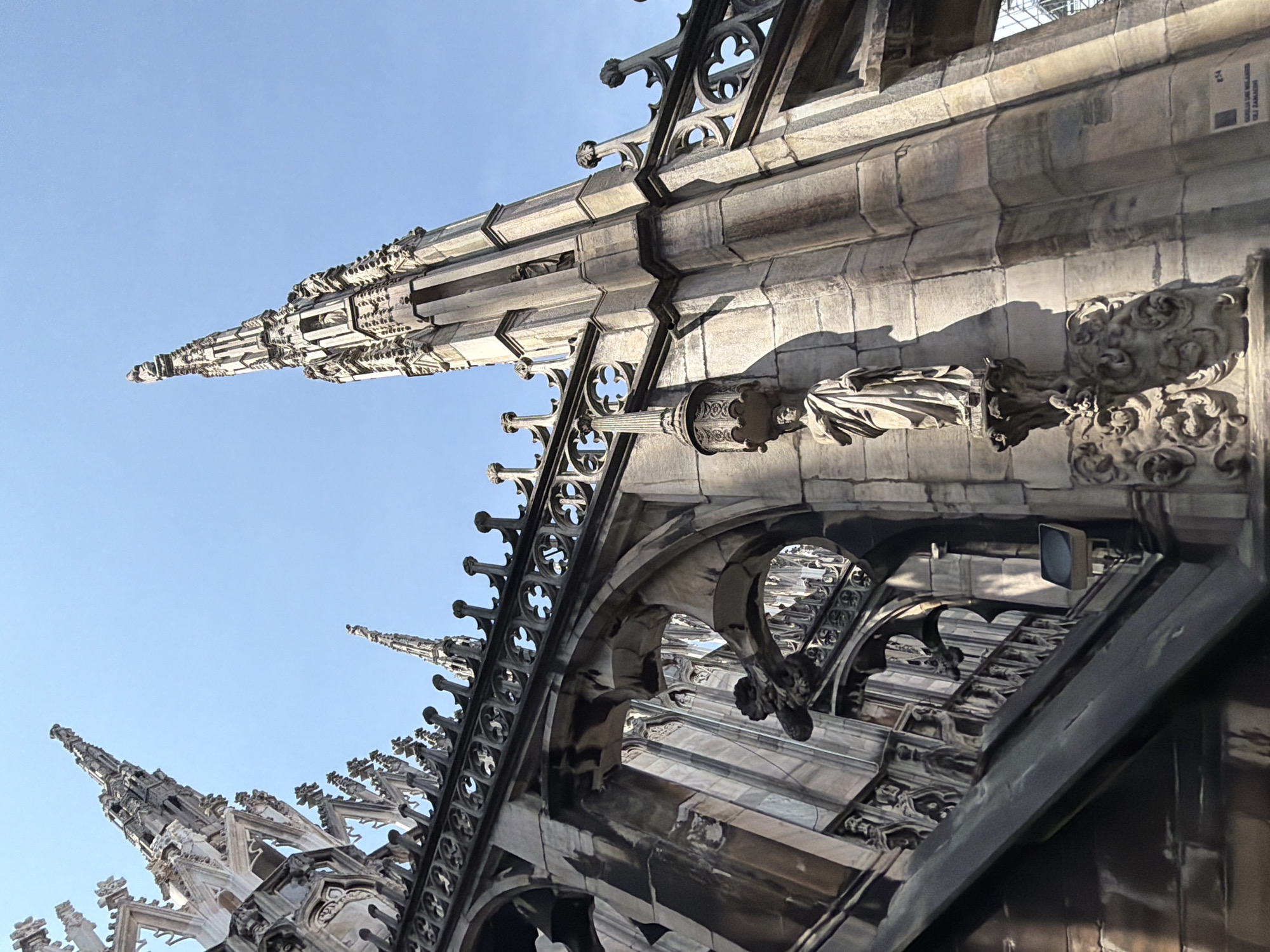 The image captures a close-up, low-angle view of the architectural details of the Duomo di Milano (Milan Cathedral) in Milan, Italy. The frame is filled with the cathedral's intricate Gothic spires, ornate stone carvings, and flying buttresses against a clear, light blue sky. Architectural elements like pinnacles, statues, and detailed tracery are visible, emphasizing the complexity of the cathedral's design. Sunlight creates shadows that highlight the textures and depth of the stonework. The architectural style is definitively Gothic, typical of the Duomo.