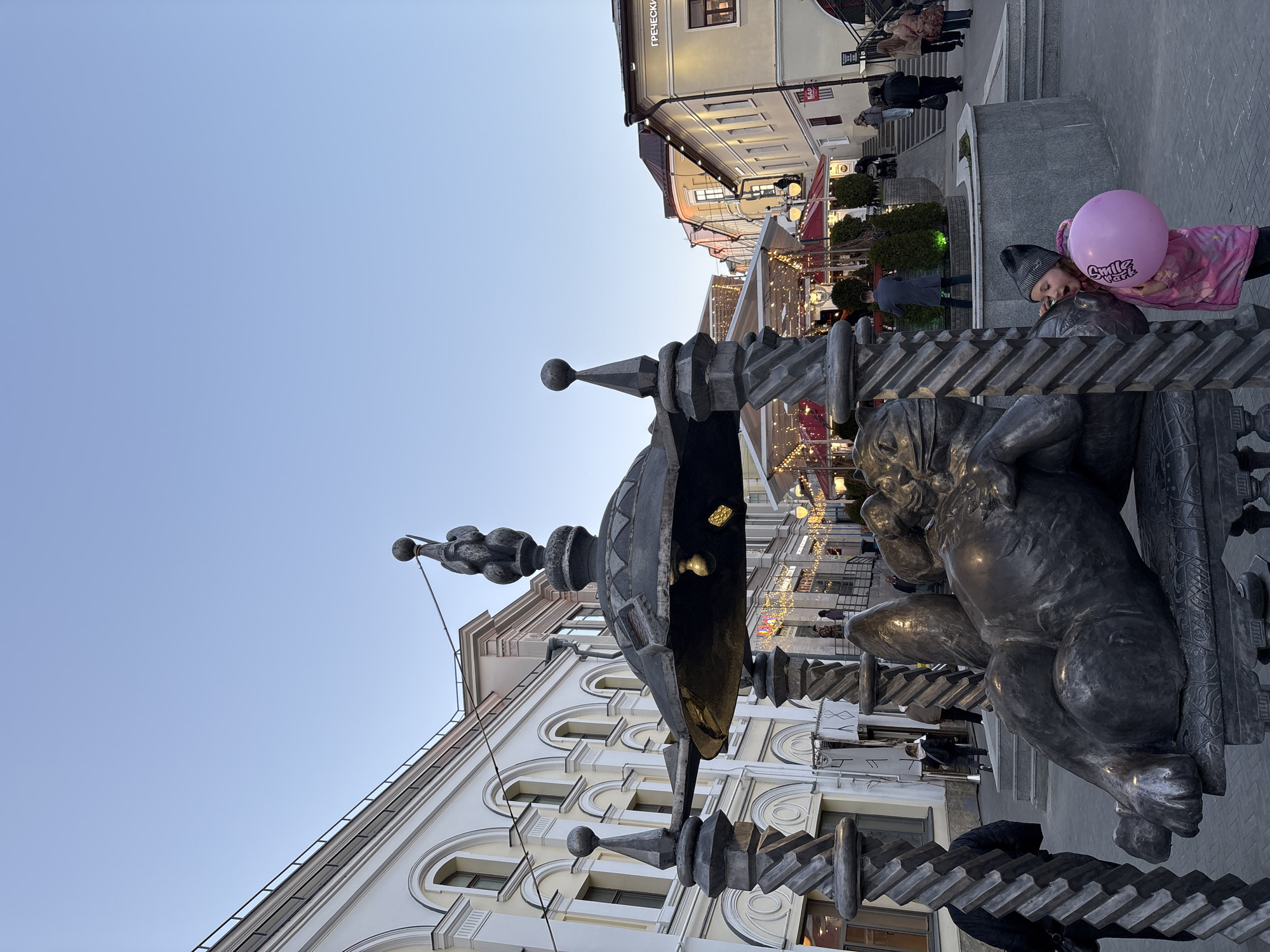 The image shows a sculpture in Kazan, Russia, with a girl holding a pink balloon next to it. The sculpture features a decorative metal structure with ornate details and figures, possibly of lions. The girl is standing beside the sculpture, partially leaning on it with the balloon in her hands. Behind the sculpture and the girl, there is a street scene with buildings, some of which are adorned with lights, suggesting a festive or evening atmosphere. People can be seen walking in the background. The sky is clear, indicating it is either early morning or late afternoon. The buildings have a classic architectural style with arched windows and white facades.
