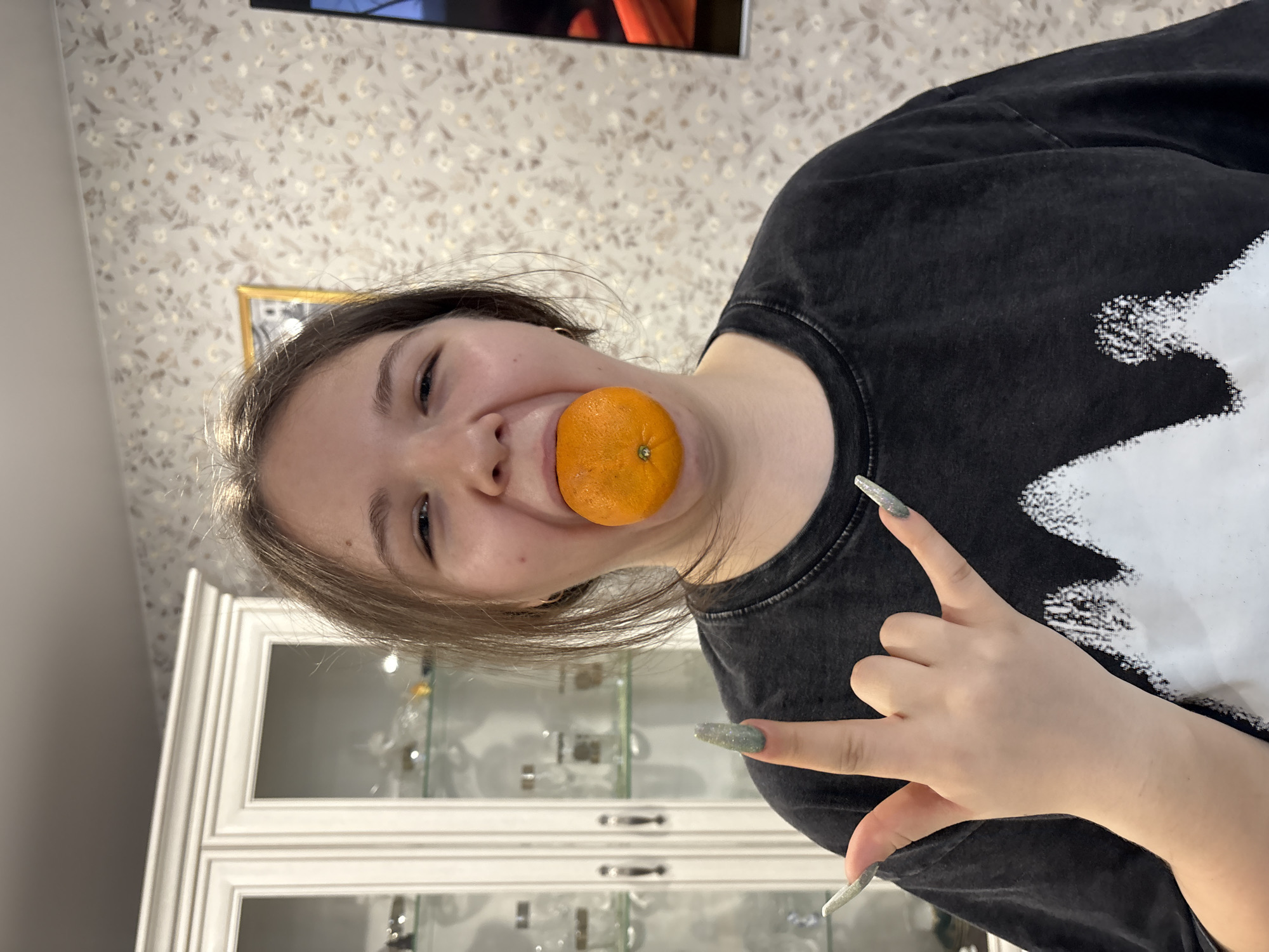 A young woman holds an orange in her mouth, smiling slightly, while gesturing with her left hand. She is wearing a dark grey t-shirt with a white design. The background includes a white cabinet with glass doors, a wall with patterned wallpaper, and what appears to be a framed picture hanging on the wall. While it's not possible to confirm the location in the city of Chaikovsky, Russia, based on the visual information provided, the interior setting suggests a domestic environment.