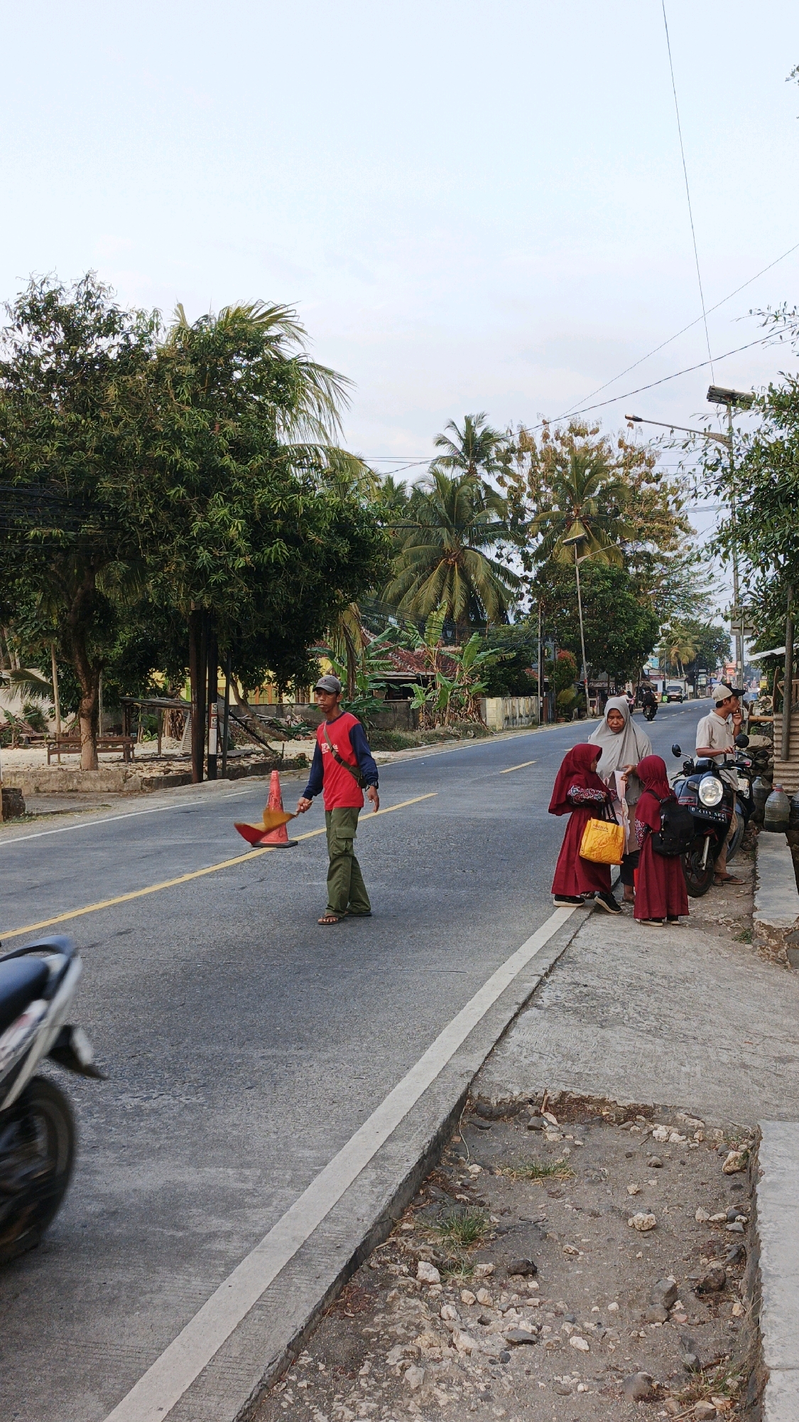 In the city of Cibenda, Indonesia, a man wearing a red shirt, a dark undershirt, a cap, and green pants stands on the asphalt road, holding a red and orange traffic cone and a yellow flag. He is positioned on the left side of the road, near a yellow line. Behind him, palm trees and lush green foliage line the road, with some houses visible in the background.
On the right side of the road, a concrete sidewalk runs parallel to the asphalt. A group of people are gathered on the sidewalk. A woman wearing a light grey hijab and a woman in a dark red hijab are interacting with two young girls, also in red hijabs. One of the girls is holding a yellow bag. To the right of this group, a man wearing a grey shirt and a cap sits on a motorcycle, facing the camera. Another motorcycle is partially visible in the foreground on the left side of the image, moving away from the camera. The sky is overcast, and the overall lighting suggests it is late afternoon or early morning. The environment appears to be a semi-rural or suburban area with moderate vegetation and a paved road.