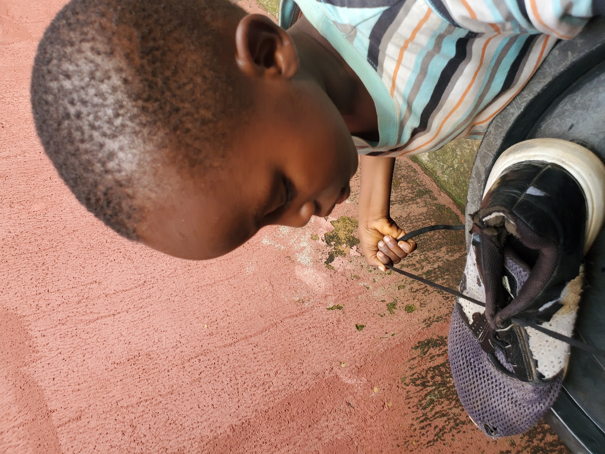 An overhead, close-up shot shows a young Black child, possibly a boy, wearing a horizontally striped t-shirt with bands of light blue, white, black, and orange. The child is focused on tying the shoelace of a worn, dark-colored athletic shoe that rests on the edge of a black plastic container. The shoe is predominantly black with white accents and a white sole, showing signs of significant use. The child's right hand is visible, holding the shoelace. The background features a textured, reddish-pink wall with patches of peeling paint and green moss, typical of outdoor structures in humid climates. The lighting suggests it is daytime. The scene appears to be an outdoor setting, possibly in a residential area. While specific details about the city of Jos, Nigeria cannot be definitively confirmed from the image alone, the presence of such a wall texture and environmental conditions could be consistent with urban or semi-urban areas in Nigeria.