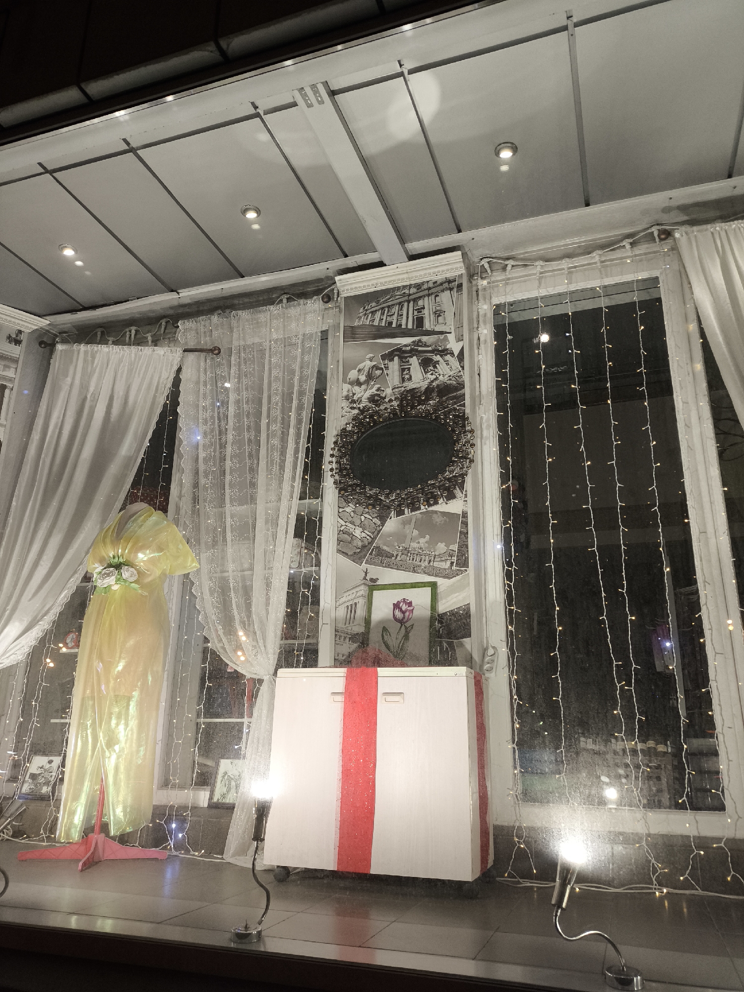 The image depicts a store window display in Moscow, Russia. A yellow dress form with a bouquet of flowers stands behind a white cabinet with a red ribbon. Behind these items are a curtain, a window covered in string lights, and pictures of Russian landmarks.