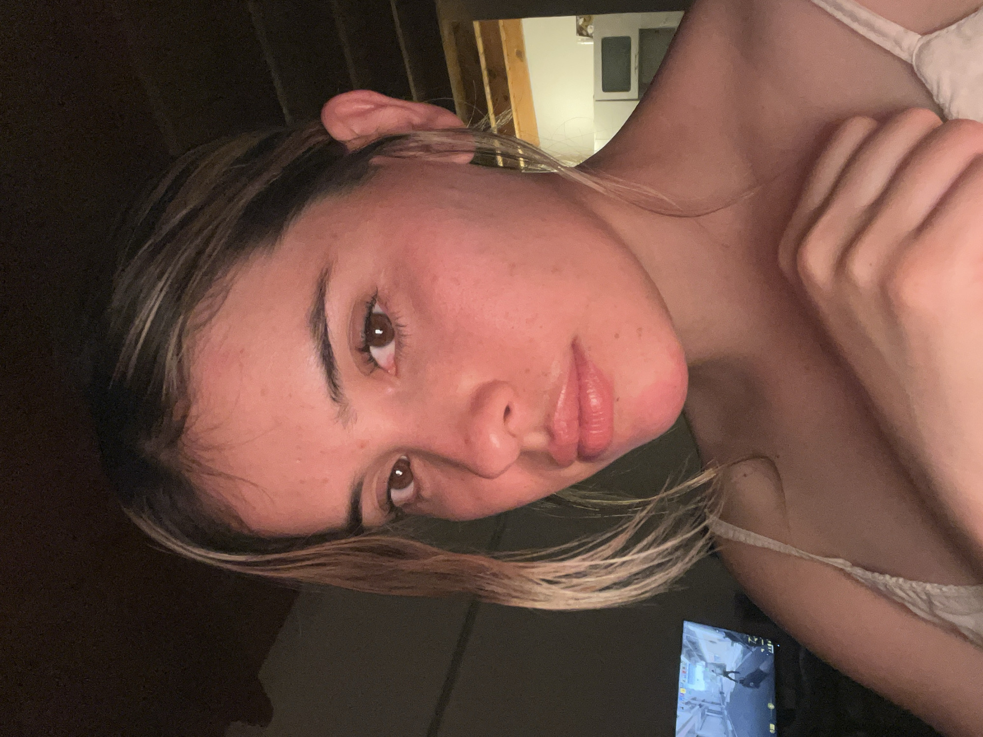 A young woman is shown in a close-up, self-portrait style image. Her face is illuminated by warm lighting, highlighting her brown eyes, light brown hair with blonde highlights, and rosy cheeks. She is wearing a white camisole. Her right hand is positioned on her collarbone, and her left arm is not visible. In the background, a television screen displays a video game, and there appears to be a cluttered room with wooden beams and a white cabinet. The image does not provide any specific information about the location in the city of Sumalao, Argentina.