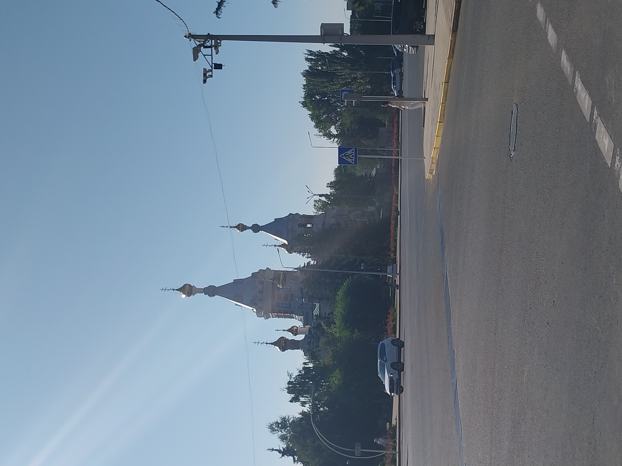 The image depicts a daytime scene in the city of Oral, Kazakhstan, showing a prominent Orthodox church with multiple golden domes. The church is situated behind a line of trees and a flowerbed. In the foreground, a light-colored SUV is turning at an intersection. A pedestrian crossing sign and a lamppost with surveillance cameras are visible on the right side of the road. The sky is clear and blue.
