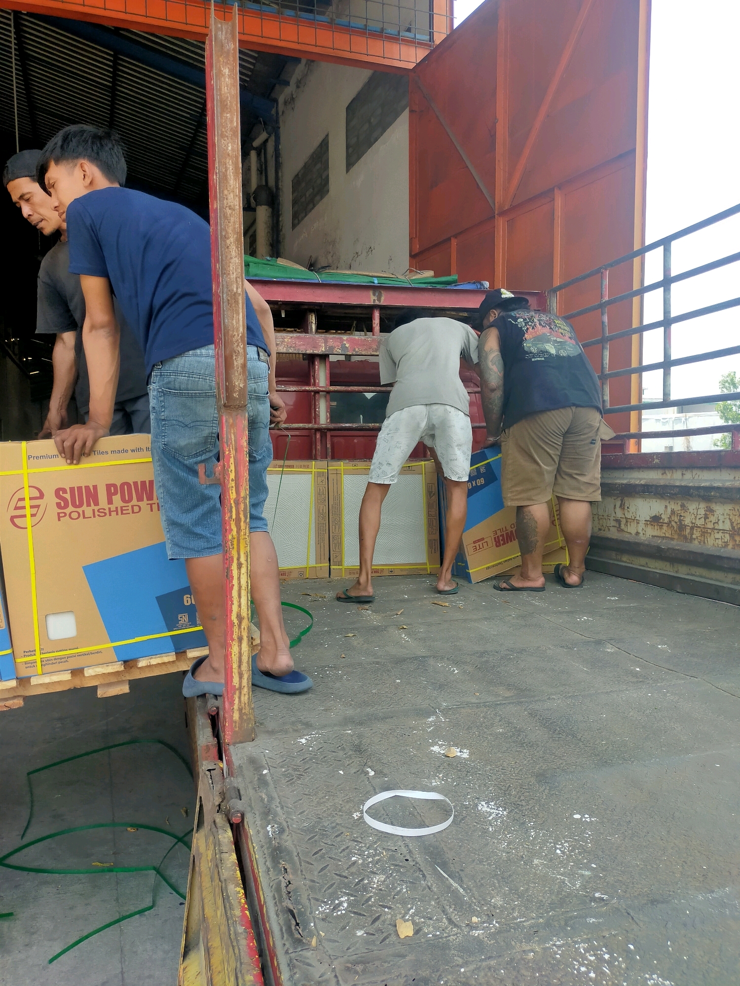 In Mlaten, Indonesia, four men are seen on the bed of a truck, engaged in loading or unloading large boxes. The truck bed is open at the back, with a metal railing along the side. The ground is a grey, textured surface, littered with debris and a few green hoses. The men are dressed casually: one in a dark t-shirt and black cap, another in a blue t-shirt and denim shorts, a third in a light grey t-shirt and patterned shorts, and the fourth in a black t-shirt with a graphic design and khaki shorts. Two of the men are actively handling a large cardboard box labeled "SUN POWER POLISHED TILES". In the background, a building with a corrugated metal roof and white walls is visible, along with a large orange metal structure, possibly part of the truck's loading mechanism or a nearby building.