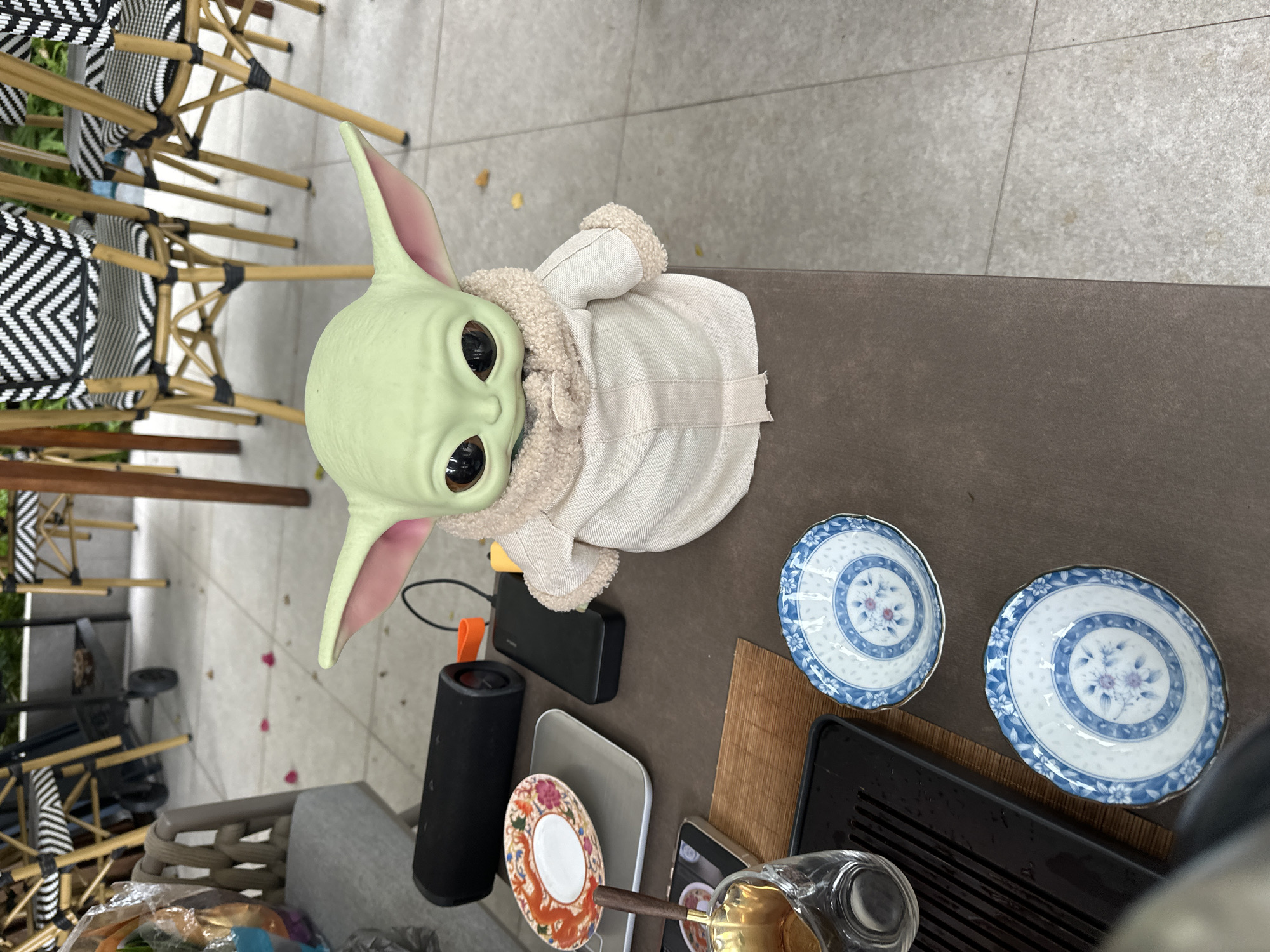 A close-up, high-angle shot shows a Grogu doll, dressed in a beige robe, seated on a brown table. To the left of the doll, a black cylindrical speaker sits next to a black power bank with an orange cable. A small, decorative plate with an orange and blue floral design is to the left of the speaker. A glass teacup with a wooden handle is in the foreground on the left. Two small, blue and white patterned bowls are positioned below the teacup. The table is on an outdoor patio with a tiled floor. In the background, several chairs with black and white patterned seats and wicker backs are visible. The scene is lit by natural light.