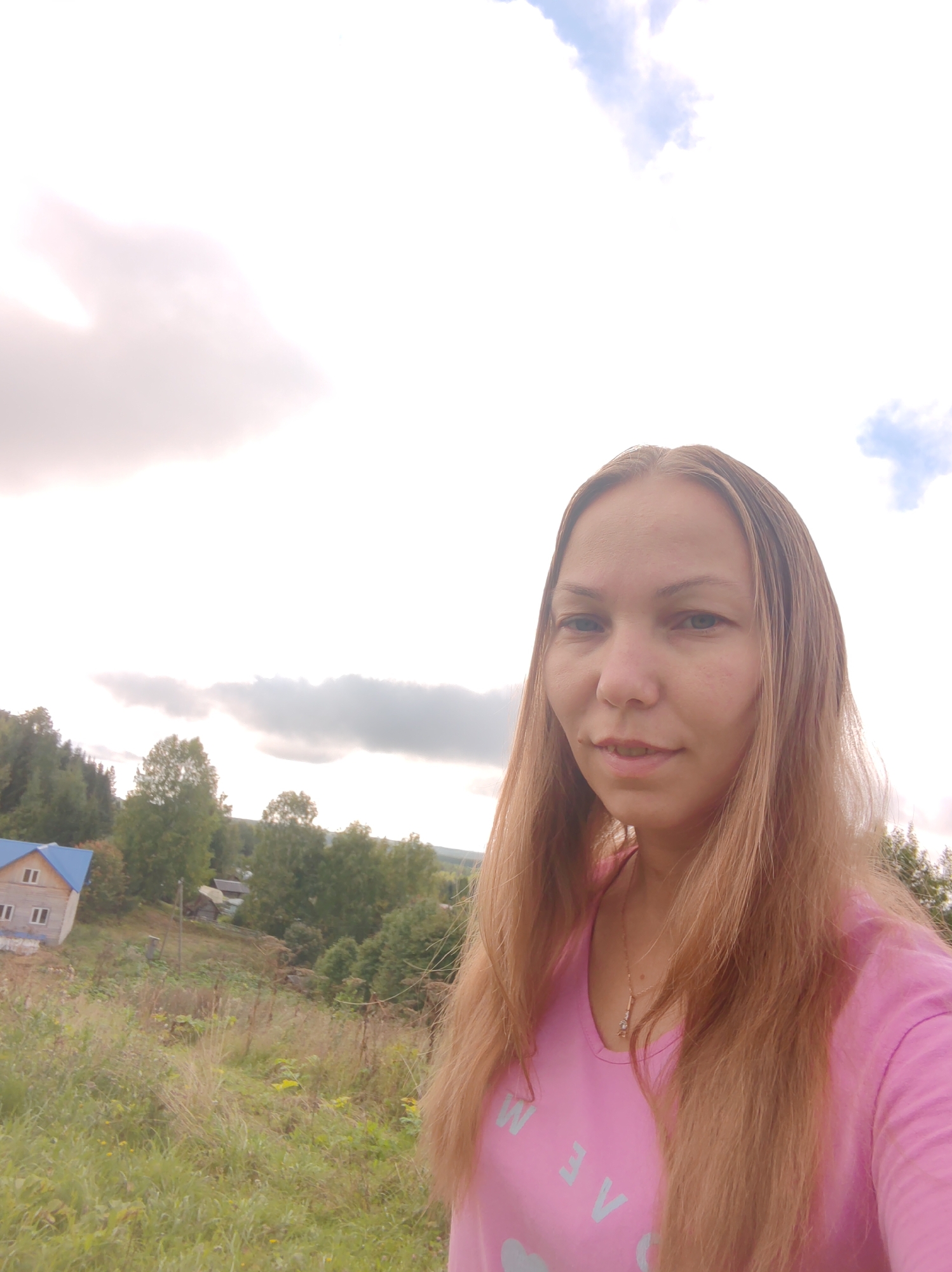 A mid-shot, outdoor, selfie-style photograph shows a woman with long, light brown hair, wearing a pink t-shirt with light blue lettering. She is looking towards the camera with a slight smile. Her facial features include light-colored eyes and fair skin with some freckles. She is wearing a necklace with a pendant.
Behind her, the environment is a rural landscape. There are rolling hills covered with green grass and wildflowers. In the middle ground, a small wooden house with a blue roof is visible on the left side of the frame, nestled amongst green trees. Further in the background, more trees and some smaller structures are scattered across the landscape. The sky is overcast with large, light grey clouds, and some patches of blue sky are visible. The overall impression is a serene, natural setting.