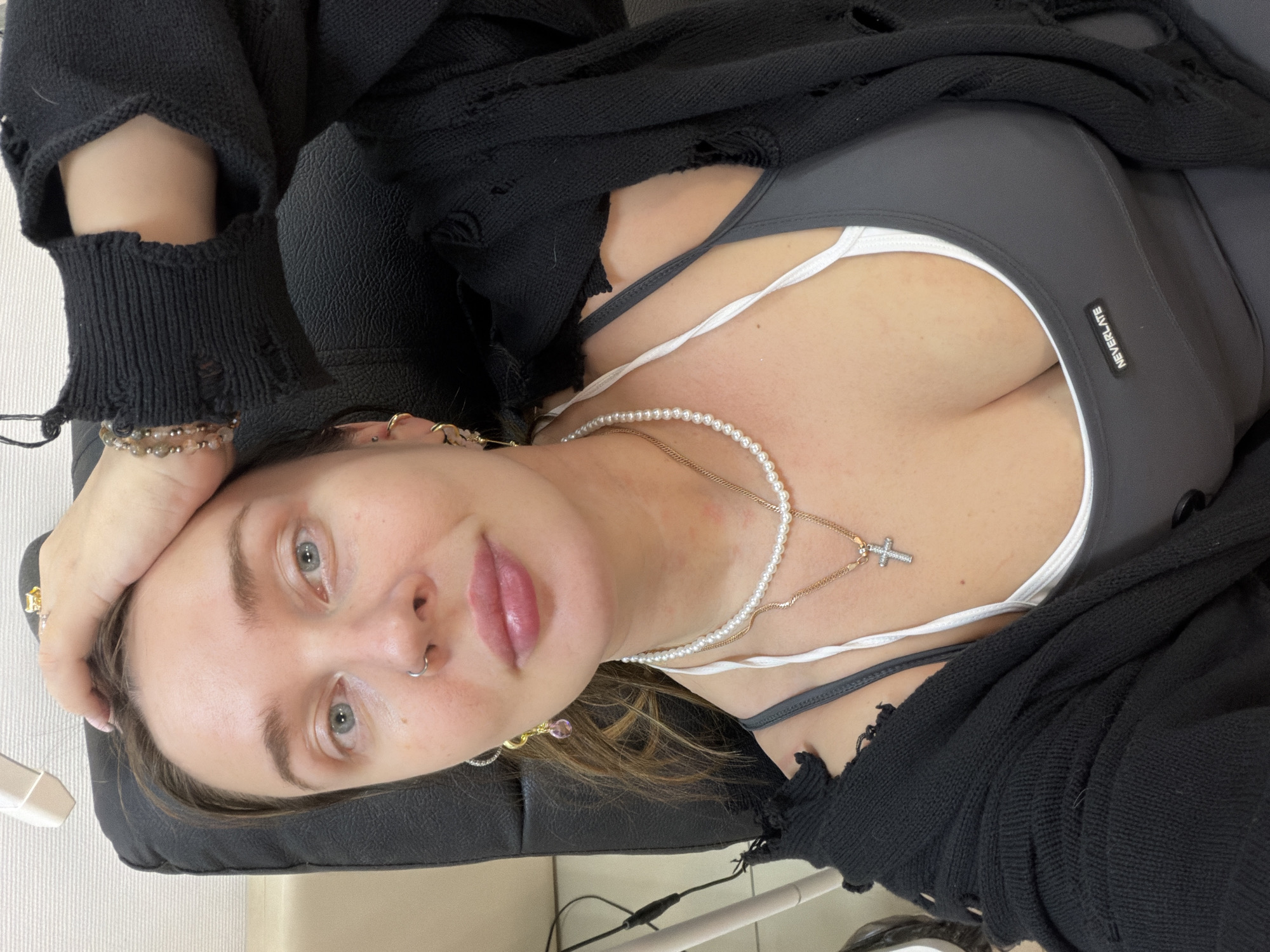 The image captures a female subject, appearing to be in her 20s or 30s, in an indoor setting described as the city of Chaikovsky, Russia. The subject is positioned in a reclining manner, resting against a black padded surface, likely a chair or examination bed, and is looking directly at the camera, suggesting a selfie. Her right hand is placed on her head, fingers slightly spread.
She has light-colored eyes and a silver hoop nose ring in her left nostril. Her lips appear full, with a pinkish tint. Reddened areas are visible on her neck and upper chest. She is wearing a black, distressed knit cardigan or sweater, worn open. Underneath, she has on a grey athletic-style top, possibly a sports bra, with a small black tag bearing the word "NEVERLATE" on the chest. A white strap of an undergarment is visible beneath the grey top.
Jewelry includes a multi-strand beaded bracelet on her right wrist. Around her neck, she wears a string of white pearls and a separate gold-colored chain with a small, stone-studded silver-colored cross pendant. Multiple piercings are visible on her ears, with a gold-colored hoop earring featuring a purple dangle in her right ear.
The background shows a light-colored wall section to the left, partially obscured by a white fixture or equipment piece. Wires are visible along the left side of the frame. The remainder of the immediate background is the dark padded upholstery of the chair.