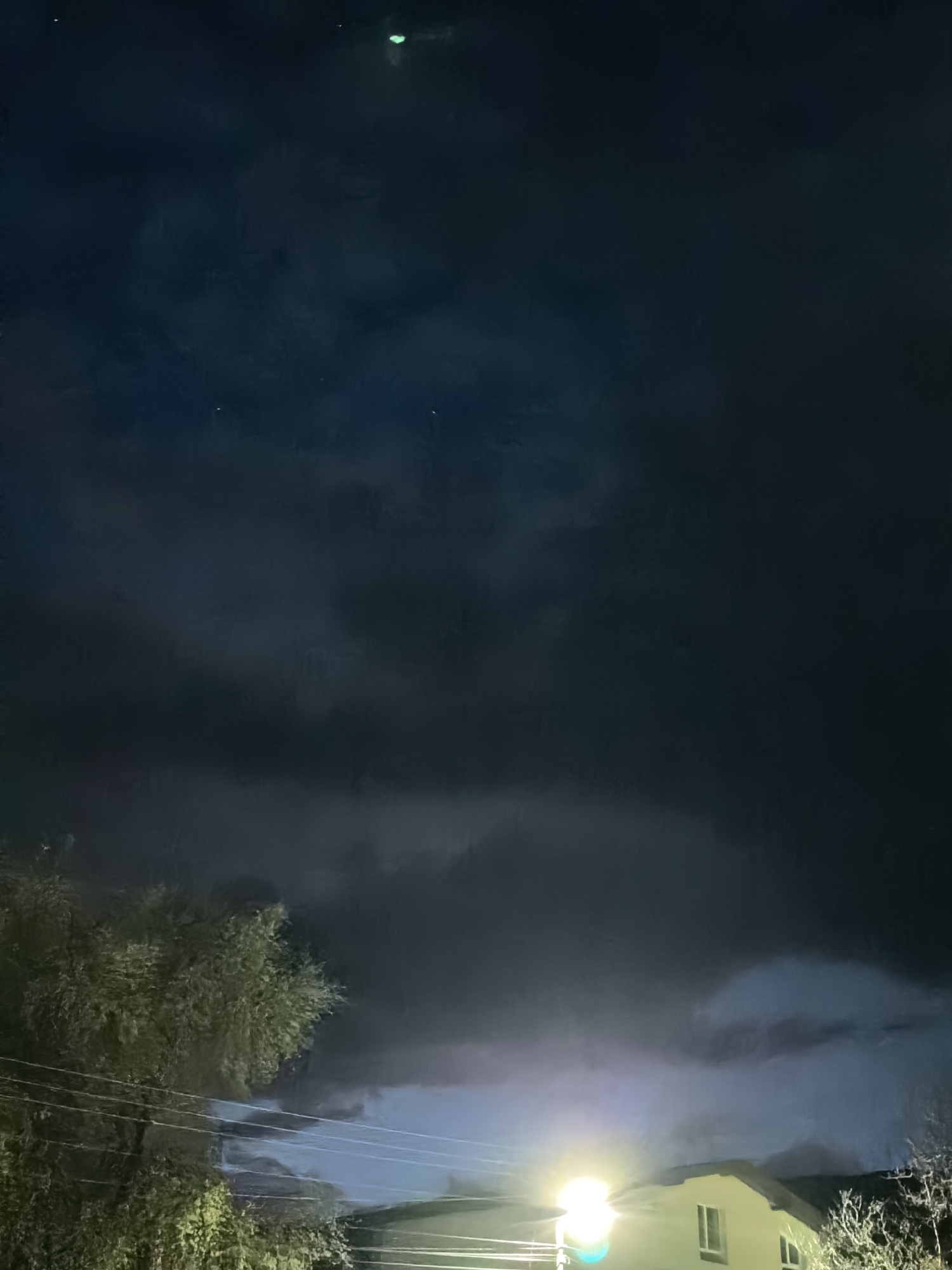 The image depicts a nighttime scene in an unknown location. The sky is dominated by dark, heavy clouds. A light source, likely a street lamp, illuminates the base of the image, highlighting a nearby house. Trees are visible on the left and right edges of the frame. Overhead power lines are seen against the sky, crossing the image. The lack of detail in the dark areas makes it difficult to discern specific features.