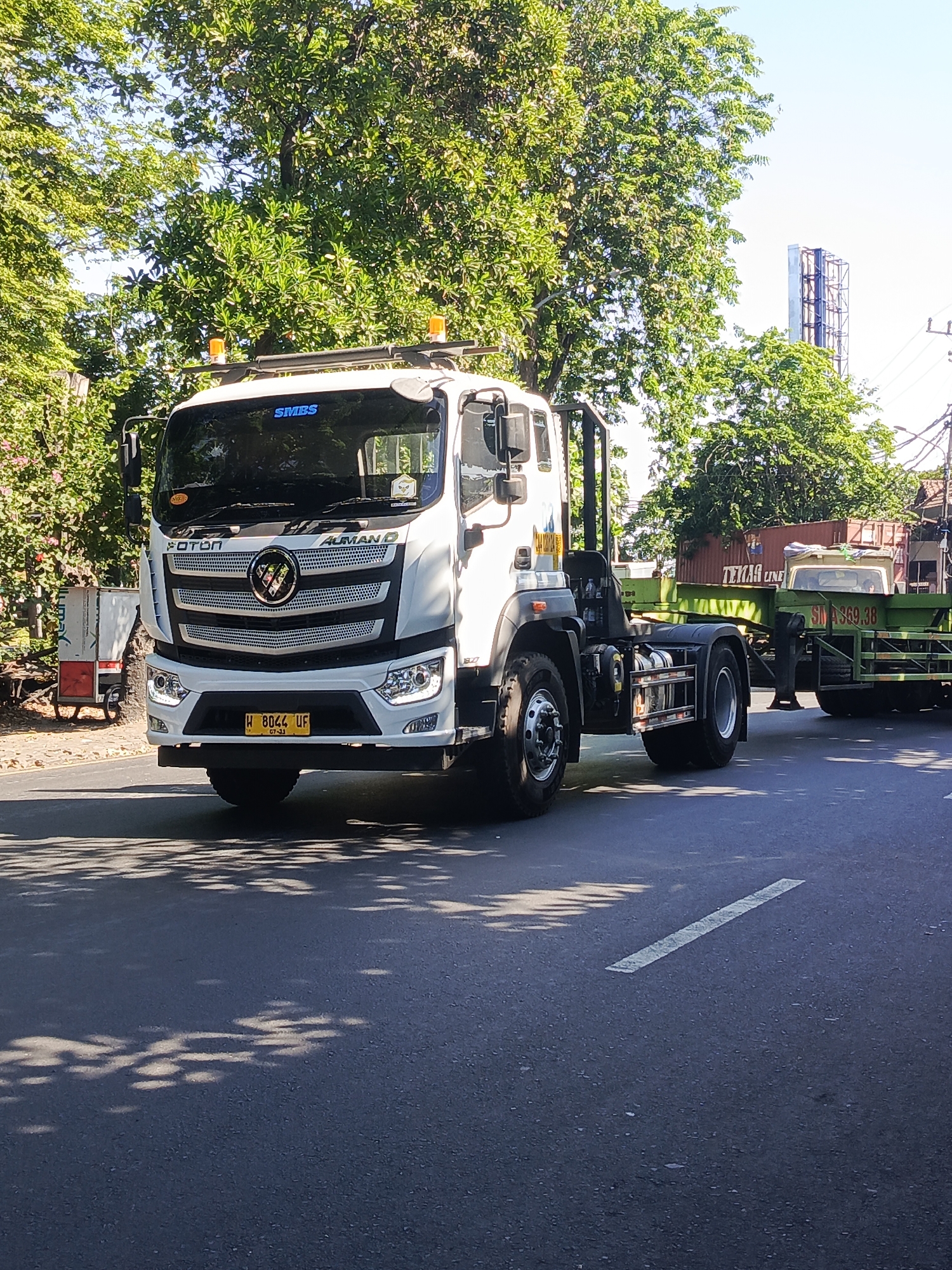 A white Foton Auman truck with a blue "SMBS" logo on the windshield is driving on a road in Surabaya, Indonesia. The truck is in the foreground, and a green trailer with a red shipping container is attached to it. The trailer has the text "TEKAR LINE" and "SM A369.38" visible. There are trees and buildings in the background, and the sun is shining. The license plate on the truck reads "W 8044 UF". Shadows of trees are cast on the road.