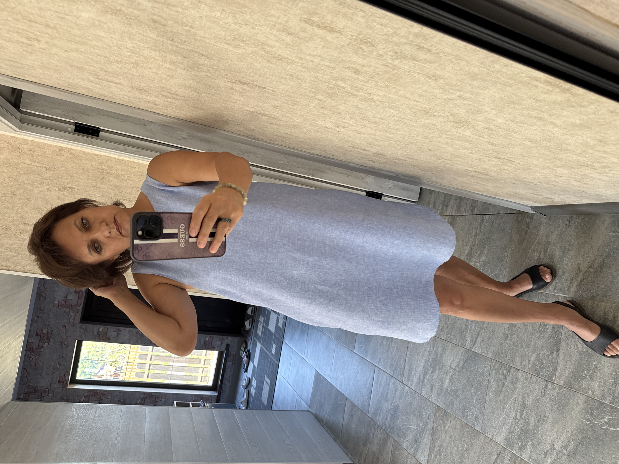 The image shows a woman in a light blue dress and black sandals taking a mirror selfie. She is standing in a hallway with tiled floors and walls. In the background, a window shows a glimpse of greenery and a fence. A pair of white and several pairs of dark slippers are visible on a mat in the distance. The woman is holding a phone with a patterned case and wearing a bracelet and a ring. The lighting is natural and appears to be daytime.