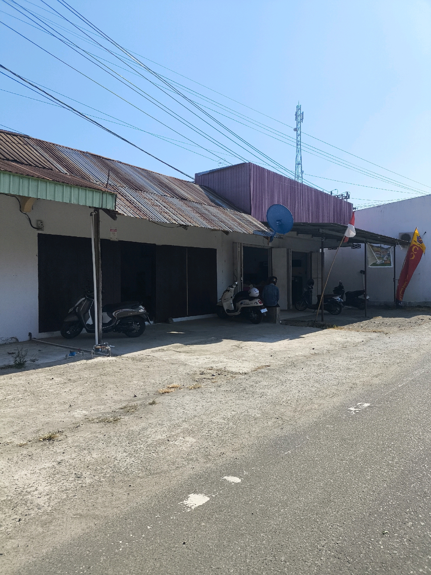 The image depicts a street view in Tilamuta, Indonesia, on a clear day. A single-story commercial building with a corrugated metal roof and a purple-colored facade is visible. The building has a covered walkway in front, with several parked motorcycles and scooters. Two individuals are present, one sitting on a bench outside the building, and another standing or walking near the entrance. Indonesian flags are displayed on poles near the entrance. A satellite dish is mounted on the roof. Electrical wires crisscross the sky above the building. The ground in front of the building is a rough, paved surface, and a road is visible in the foreground. A telecommunications tower is present in the distance.