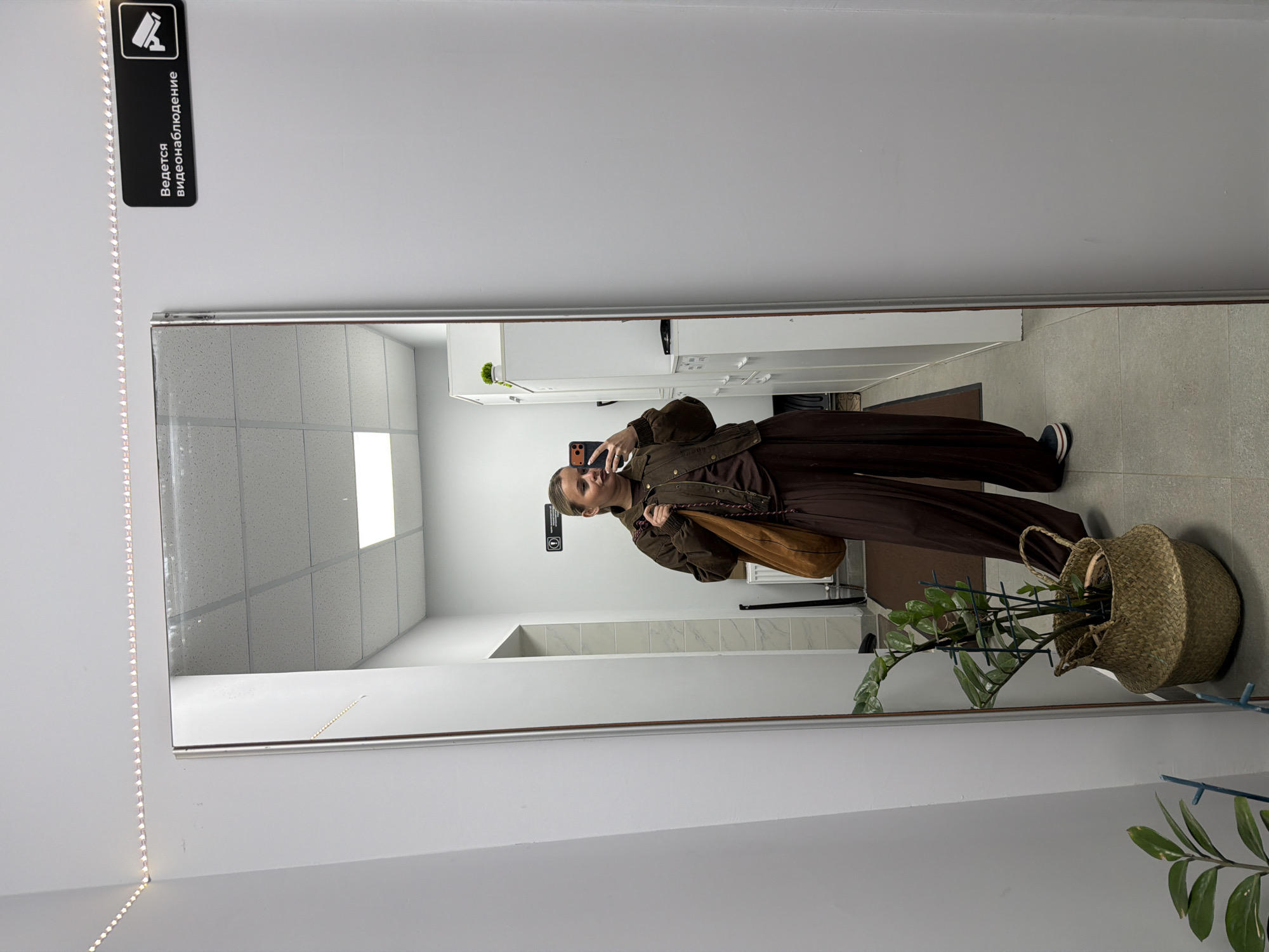 A full-length mirror reflects a woman taking a selfie in what appears to be an indoor public space. The woman is wearing a brown jacket, matching pants, and black and white sneakers. She is holding a phone in her right hand and a brown shoulder bag is slung over her left shoulder.
The reflection also reveals a sterile environment with white walls, white ceiling tiles, and light-colored floor tiles. Cabinets are visible in the background. To the right of the mirror is a straw basket containing a leafy green plant. At the top left corner of the image is a black sign with an icon and Cyrillic text, suggesting the location is likely Russia.
Given the provided location of Sarapul, Russia, this scene is likely a bathroom or utility area in a commercial or public building.