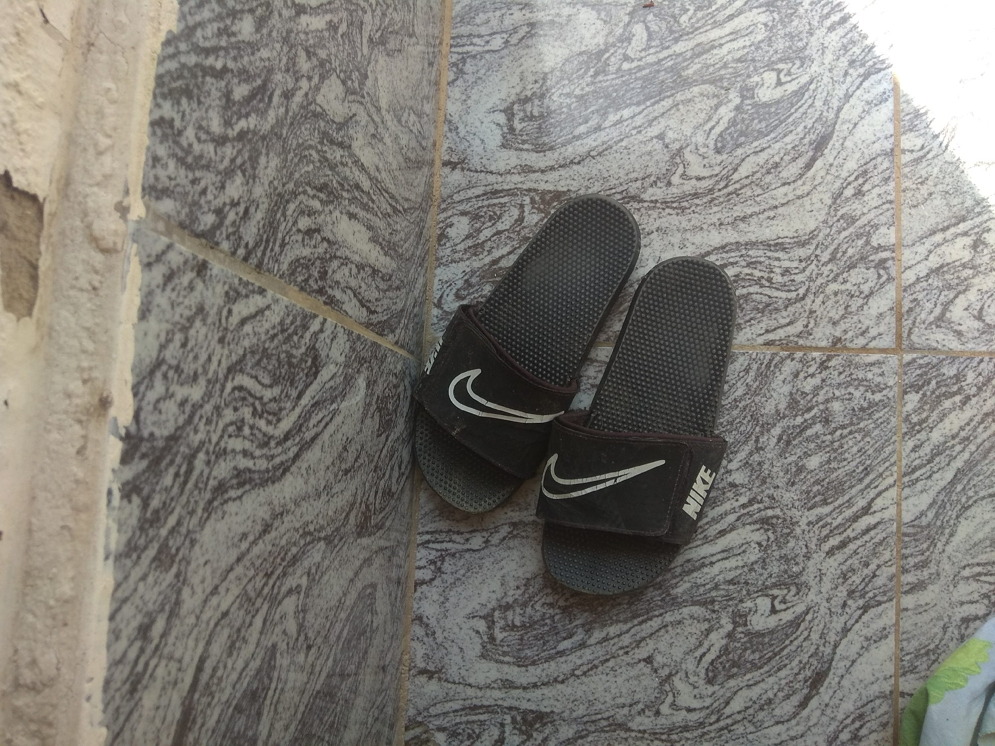 A pair of black Nike slide sandals are placed on a tiled floor in the corner of a room. The tiles have a marbled pattern in shades of gray and white. To the left of the sandals, a rough, textured wall is visible, with sections of paint peeling away. The lighting suggests it is daytime, with a bright light source on the right side of the image. The scene appears to be an outdoor or semi-outdoor space, possibly a patio or walkway, given the tiled floor and the rough wall texture, which could be part of a building exterior. The sandals are positioned with their straps facing upwards, and the Nike logo is clearly visible on both. The ground is dry and clean. The overall impression is one of a simple, utilitarian setting.