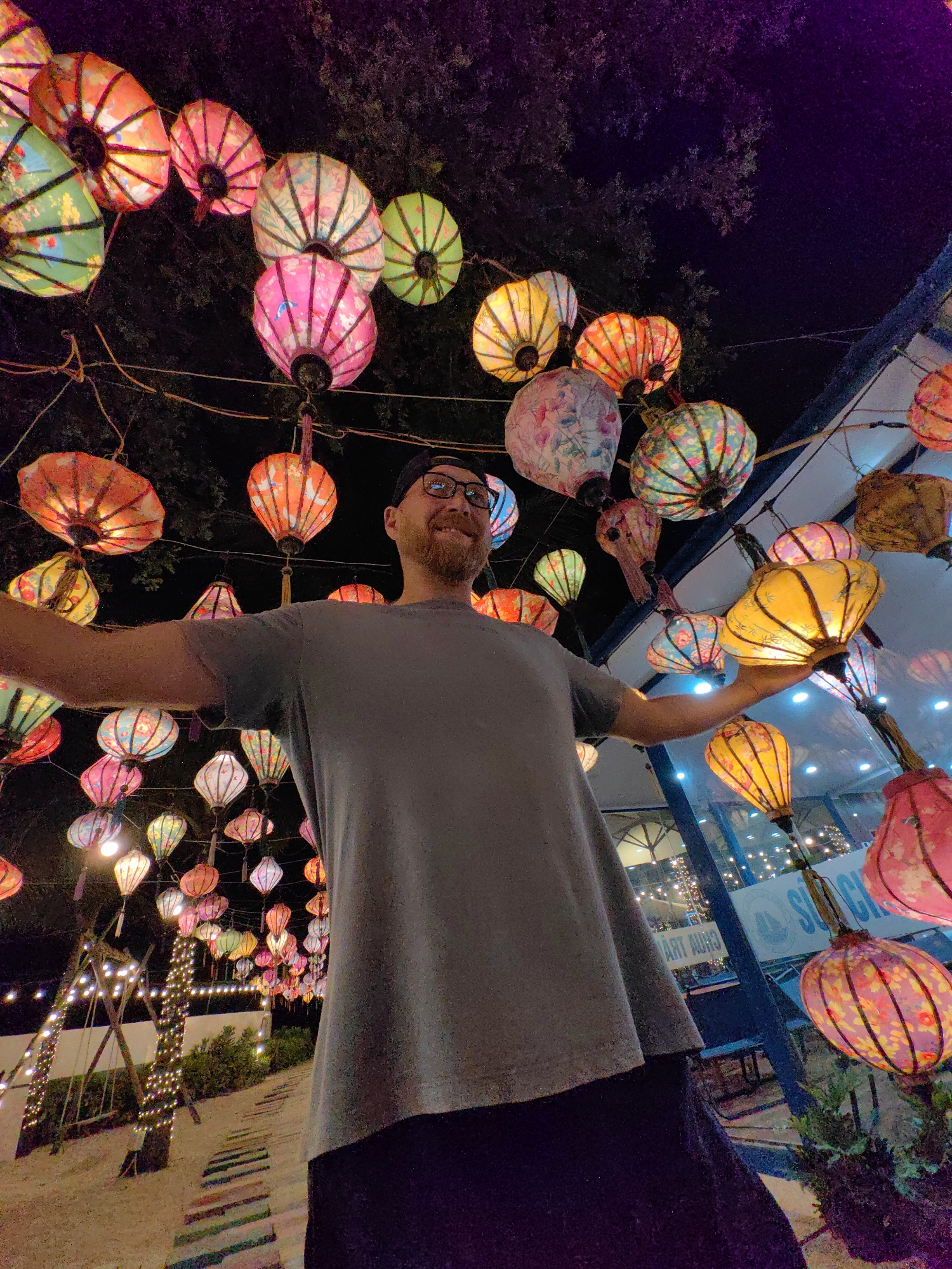 A man with glasses and a beard, wearing a gray t-shirt and a dark baseball cap, stands with his arms outstretched amid a display of colorful lanterns in Bai Chay Ward, Vietnam. The lanterns are of various colors and patterns, strung overhead and to the side, illuminating the scene at night. In the background, there are trees and a building with lights visible through the glass. The ground appears to be a pathway with colored segments.
