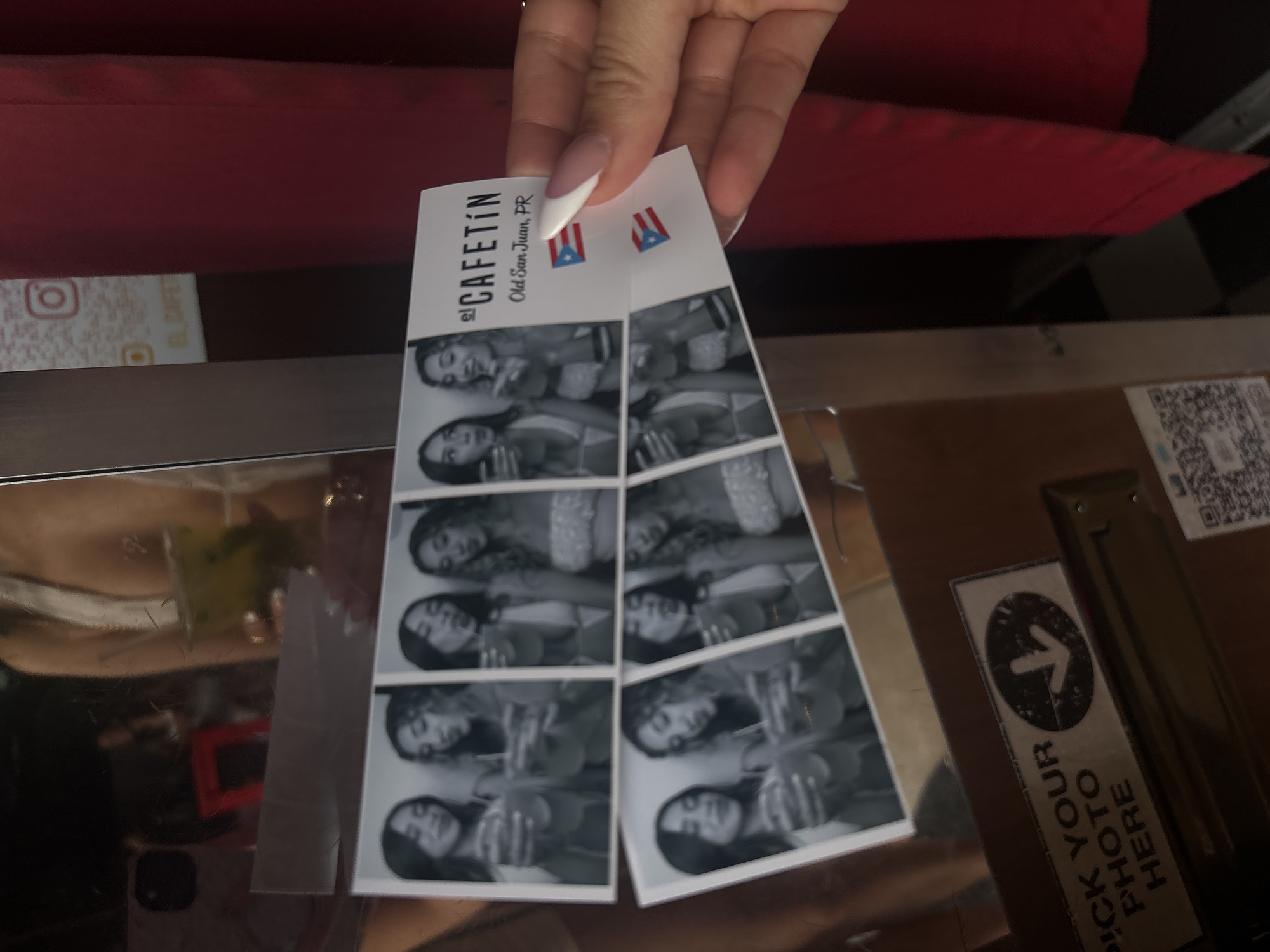 A black and white photo booth strip is held by a person's hand, displaying four rows of images of two young women. The women are posing with drinks and appear to be in a casual, social setting. The strip also features the name "el CAFSTIN" and "Old San Juan, PR" along with two Puerto Rican flags. In the background, a QR code and a sign that reads "PICK YOUR PHOTO HERE" are visible, suggesting the photo booth is in a public establishment. The overall environment appears to be indoors, possibly a bar or entertainment venue, with red fabric visible in the background.