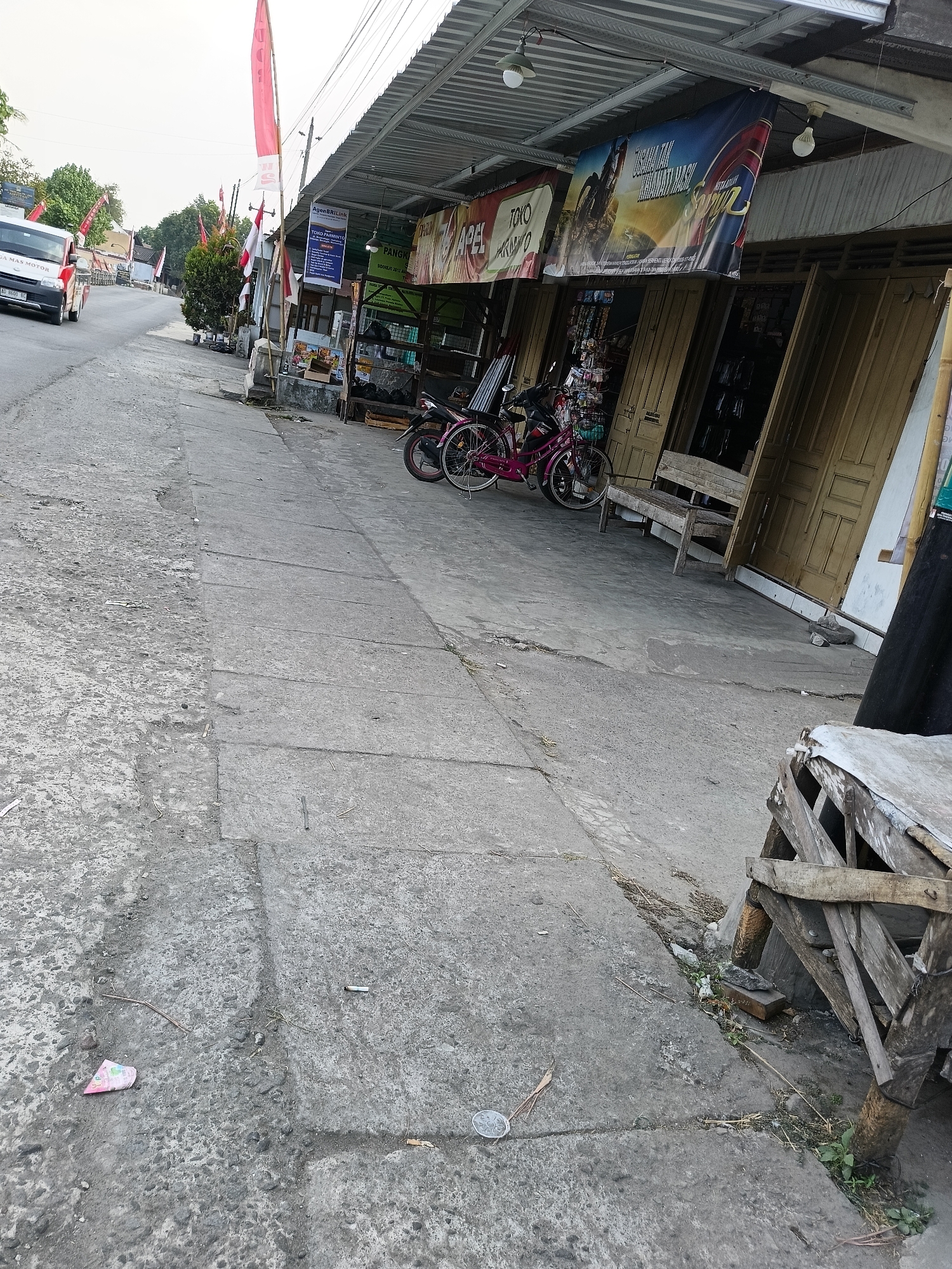 The image depicts a street scene in Sukorini, Indonesia. On the left side of the road, a white and red van is traveling away from the camera. The road is paved with asphalt and concrete sections. A sidewalk, also made of concrete, runs parallel to the road. Along the sidewalk, there are several shops with their doors open. These shops have banners displayed in front of them, advertising various products and services. One banner features a motorcycle rider, while another shows text in Indonesian. Red and white flags are also visible, suggesting a national celebration or event. Two bicycles, one pink and one dark-colored, are parked on the sidewalk in front of one of the shops. A wooden bench is placed in front of another shop. The environment appears to be somewhat cluttered, with debris and small objects scattered on the ground, including a pink piece of paper, a cigarette butt, and a coin. The buildings are simple structures with corrugated metal roofs. The lighting suggests it is daytime, possibly late afternoon, given the soft light. The scene is typical of a commercial area in a small Indonesian town.