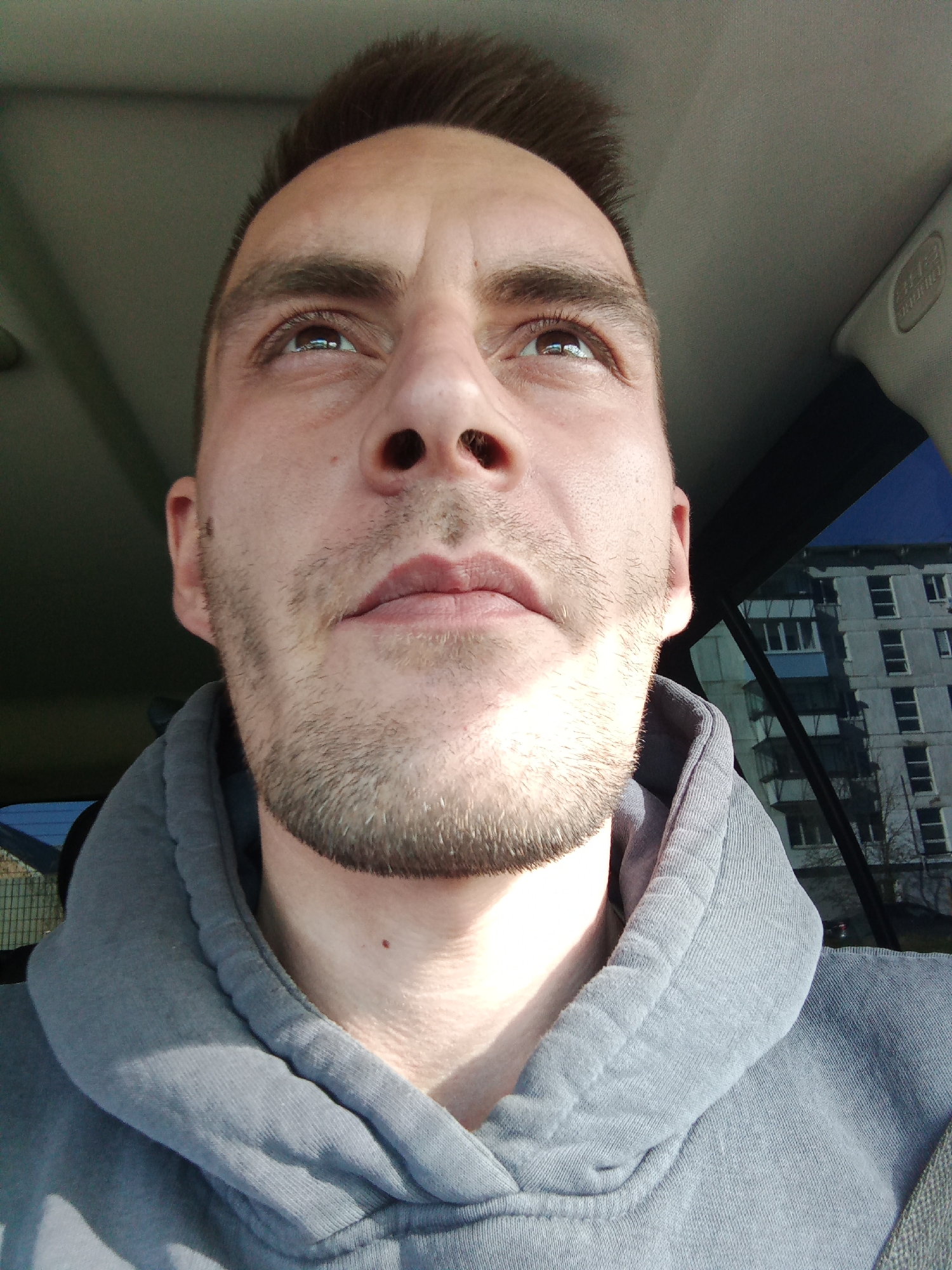 A close-up, upward-angled shot of a light-skinned man with short brown hair and stubble, wearing a grey hooded sweatshirt. The man is looking upwards and slightly to the right. The background features the interior of a vehicle, indicated by the headliner and window. Through the window, a multi-story residential building is visible. The style of the building suggests a location in a Russian city; considering the prompt, a possible location could be Chaikovsky, Russia.