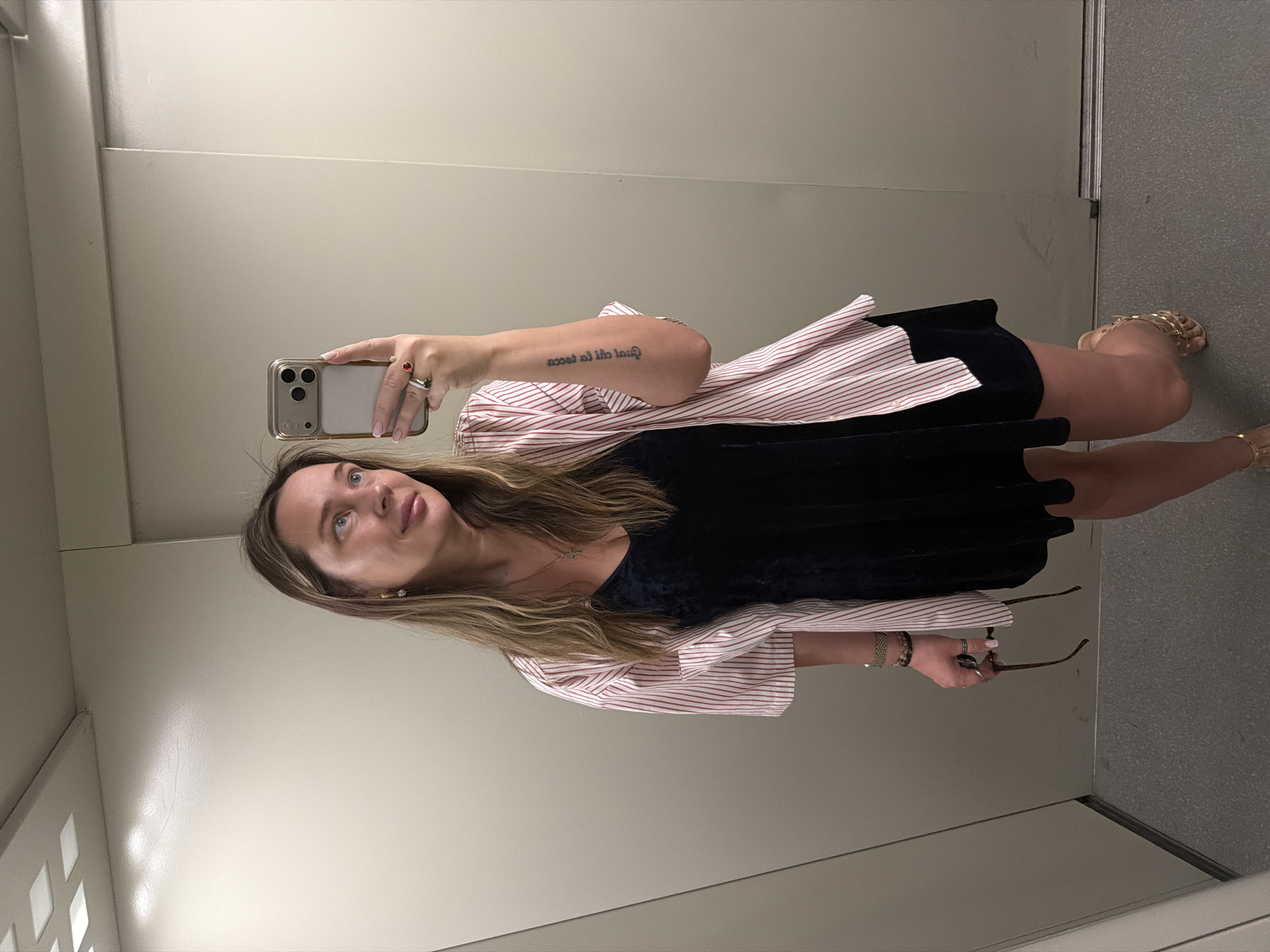 A woman stands inside an elevator in Chaikovsky, Russia. She is taking a selfie with her smartphone. The woman has medium-length blonde hair, fair skin, and blue eyes. She is wearing a black dress layered over a long-sleeved shirt with red and white stripes. She wears multiple rings on her fingers and bracelets on her wrists. She is wearing gold-colored sandals. She holds a pair of eyeglasses in her left hand. The elevator has plain light-colored walls and a gray floor.
