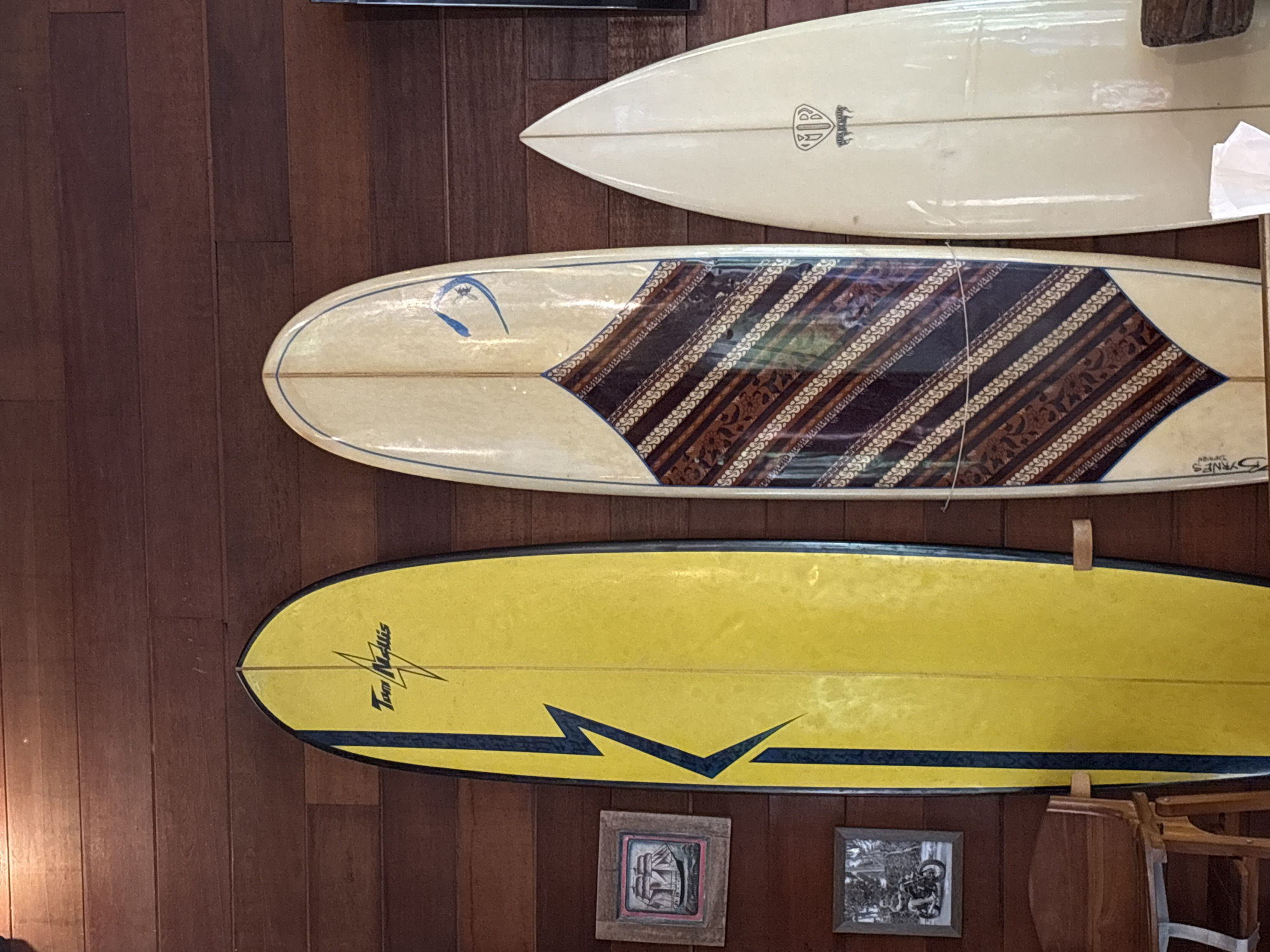Three surfboards are displayed vertically on a dark, reddish-brown wooden paneled wall, likely indoors in Pekutatan, Indonesia.
The top surfboard is a light cream color with a pointed nose. A black logo featuring a stylized "D" and possibly "Dolphin" is visible towards the nose.
The middle surfboard is also light cream but features an intricate diagonal pattern across its body in shades of brown, black, and cream, resembling a batik or tribal design. A blue, abstract, bird-like or wave-like emblem is near its nose. The text "BYRNES Design" is faintly visible near the tail on the right side.
The bottom surfboard is predominantly bright yellow with black outlines along its edges. A distinctive black lightning-bolt-like graphic runs along its lower half. The name "Tom Nellius" is written in black script near the nose. This surfboard is held by two visible wooden mounts.
Below the bottom surfboard, two small framed pictures are hung on the wall. The left frame contains an abstract, grayscale drawing of a structure. The right frame shows what appears to be a vintage black and white photograph, possibly depicting people on motorcycles. To the far right, part of a wooden chair or stool is visible. No activity is occurring; the surfboards and pictures are decorative elements in the indoor setting.