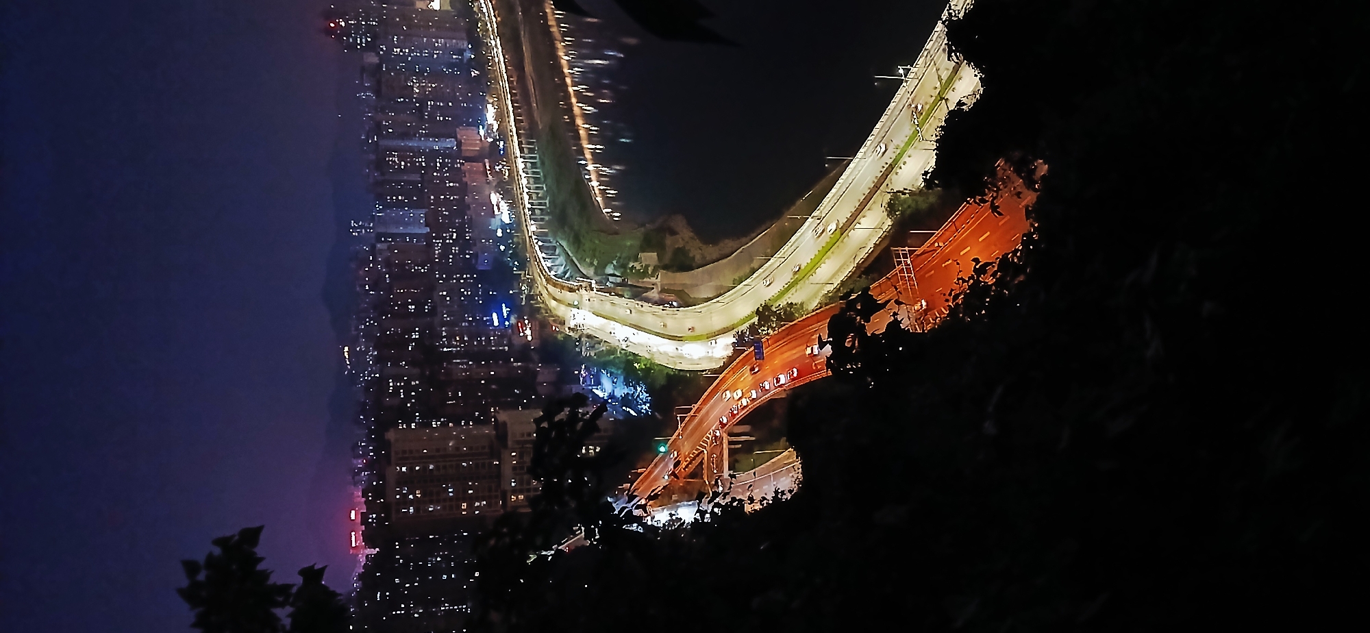 The image depicts a night view of Shapingba District, Chongqing, China, rotated counter-clockwise by 90 degrees. Prominent features include a curving elevated highway lit with white streetlights and passing vehicles. A second, lower bridge, possibly under construction or maintenance, is lit with orange lights. A city skyline with numerous lighted buildings stretches along the left side, creating a visual contrast between the urban environment and the bridge structures. The foreground is obscured by dark foliage, suggesting a vantage point from a hillside or elevated terrain. The sky is a gradient of dark blue and purple.