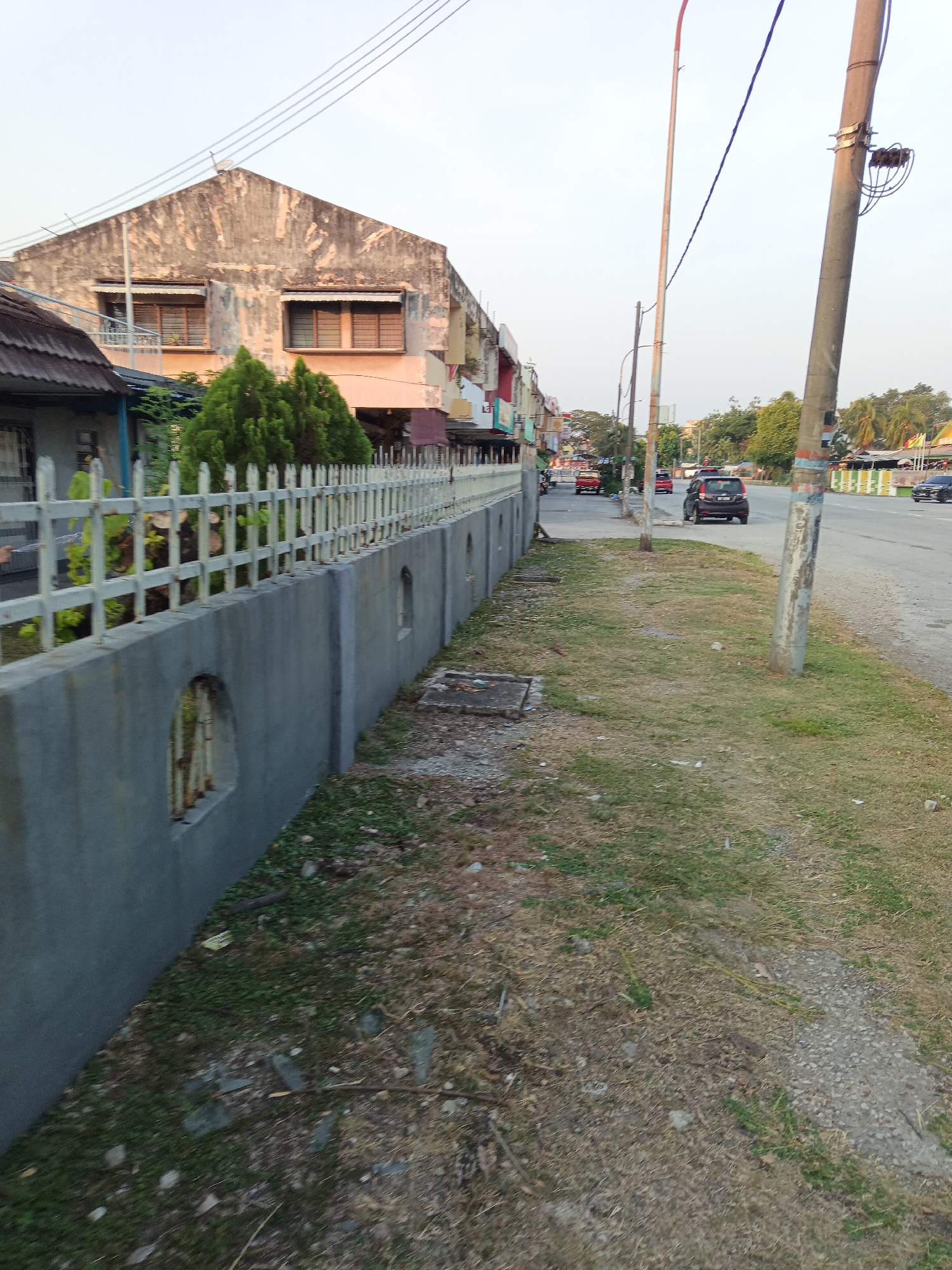 The image depicts a street scene in Klang City, Malaysia, possibly near a residential area. The perspective is from ground level, looking down a paved road lined with a low concrete wall and a white metal fence. Beyond the fence, there is a building with a weathered facade and several windows. Across the road, there are other buildings, shops with awnings, and trees. Several cars are visible on the road, including a black SUV in the foreground. Utility poles and power lines stretch across the sky. The area appears to be outdoors, with dry grass and some scattered debris on the side of the road. The lighting suggests it is daytime, possibly late afternoon due to the soft light.