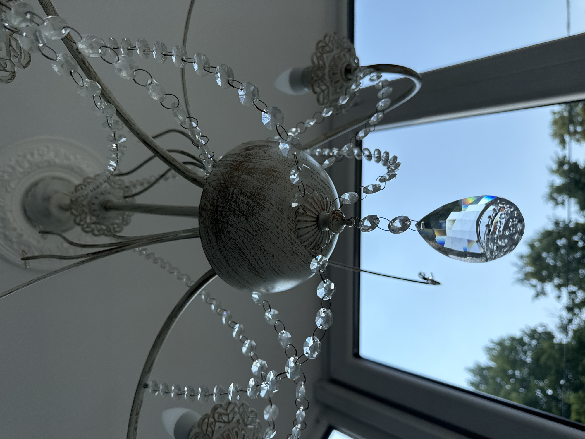 This media file shows a close-up, low-angle shot of a decorative chandelier with crystal beads and a faceted crystal pendant. The chandelier appears to be made of distressed white metal, with ornate detailing on its base and arms. Strands of clear, faceted crystal beads are strung together and hang from the chandelier arms, some cascading downwards. One large, teardrop-shaped crystal pendant is prominently featured, catching the light and showing a spectrum of colors.
The chandelier is positioned in front of a window, through which a pale blue sky and blurred green trees are visible, suggesting an outdoor scene beyond. The window frame, a dark gray color, is also visible. The lighting in the image is soft, likely natural light coming from the window, and it creates subtle reflections and highlights on the crystals.
The overall impression is one of elegant decor, possibly in a home or a decorative setting. There is no visible text. The time of day appears to be daytime, with clear to partly cloudy skies indicated by the brightness and color of the sky. The location cues of trees and sky suggest an outdoor environment seen through a window.