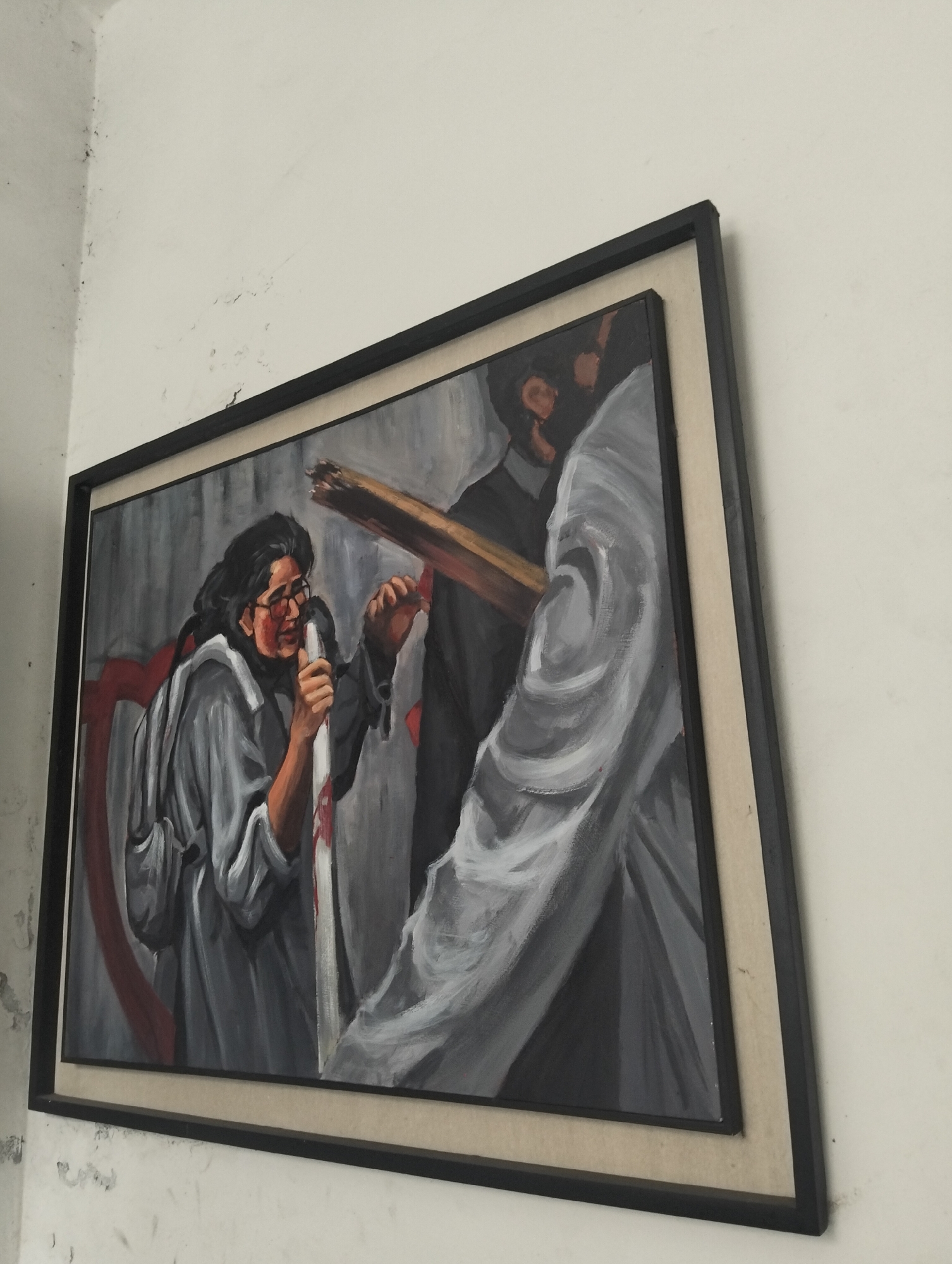 A framed painting hangs on a white wall. The painting depicts a woman with glasses holding a white pole with red markings. Behind her is a partial red heart. In front of her, a man in a dark suit carries a wooden beam, partially obscured by a white shroud. The location is Dhaka, Bangladesh.