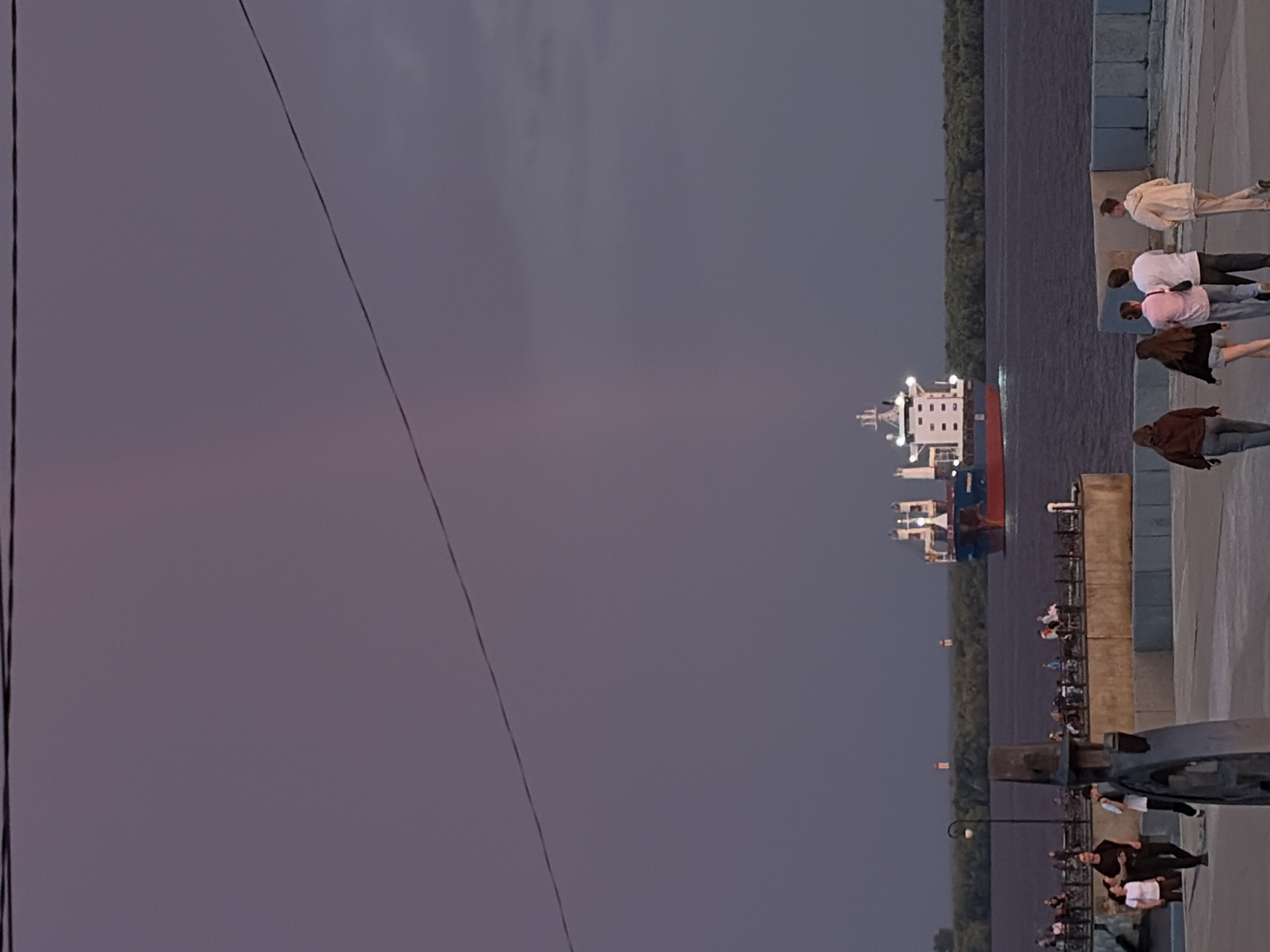 The image depicts a scene along the embankment of the Northern Dvina River in Arkhangelsk, Russia, during what appears to be twilight. A large cargo ship, painted blue and red with white superstructure, is sailing on the river, its lights illuminated. Several people are visible on the embankment, some walking and others standing. In the foreground, a dark, abstract sculpture is positioned on the paved walkway. The sky is a muted purple and grey, with a faint, vertical streak of light suggesting a rainbow or atmospheric phenomenon. The embankment itself is lined with a railing, and beyond it, a dense line of trees forms the opposite bank of the river. The overall atmosphere is calm and serene, characteristic of an evening stroll by the water.
