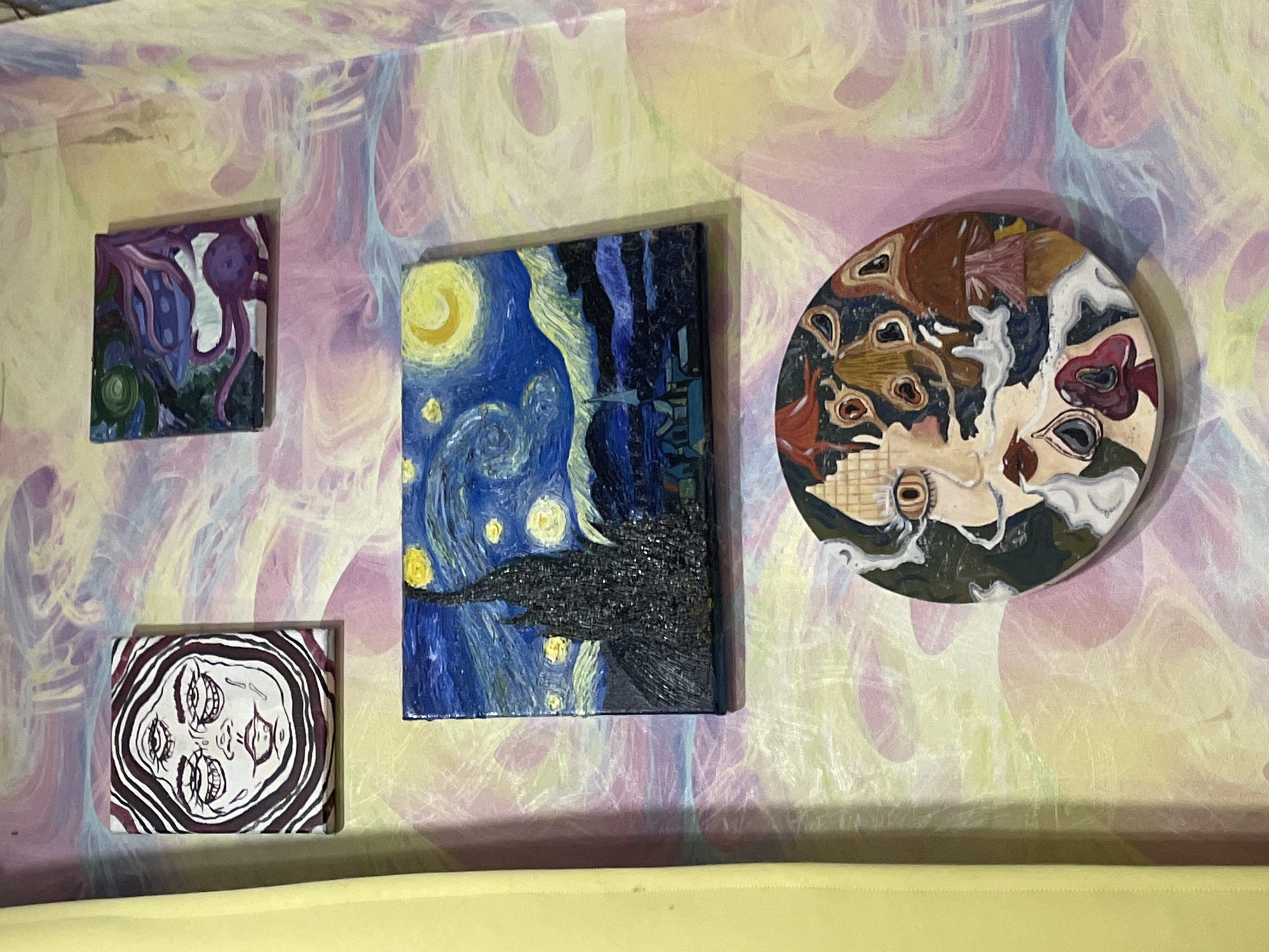 This is a close-up, eye-level, vertical shot of four small, framed paintings displayed on a wall covered in a psychedelic, swirled pattern of pastel pink, yellow, and purple. A bright yellow curtain or fabric hangs vertically on the left side of the frame, obscuring a portion of the wall.
The paintings are arranged in a staggered, somewhat haphazard fashion.
At the top left is a small rectangular painting featuring a black and white line drawing of a face with three eyes and swirling lines around the head in dark purple.
Above and to the right of the central painting is another small, square, abstract painting with purple, blue, and green colors, featuring what appears to be a stylized creature or organic shapes.
Dominating the center of the image is a rectangular painting of Vincent van Gogh's "Starry Night." It depicts a dark, jagged cypress tree in the foreground, a village with buildings and a spire in the middle ground, and a swirling night sky with a crescent moon and bright yellow stars.
Below the "Starry Night" painting is a circular painting with a surreal, psychedelic design. It features a face with large, wide eyes, red lips, and is surrounded by mushrooms and abstract shapes. The background of this painting is dark blue with scattered colorful elements.
The overall impression is of an eclectic art display in a brightly decorated, possibly bohemian or artistic, living space. The specific location within ЖК «Борисоглебское», Russia, cannot be determined from the visual information provided in the image.