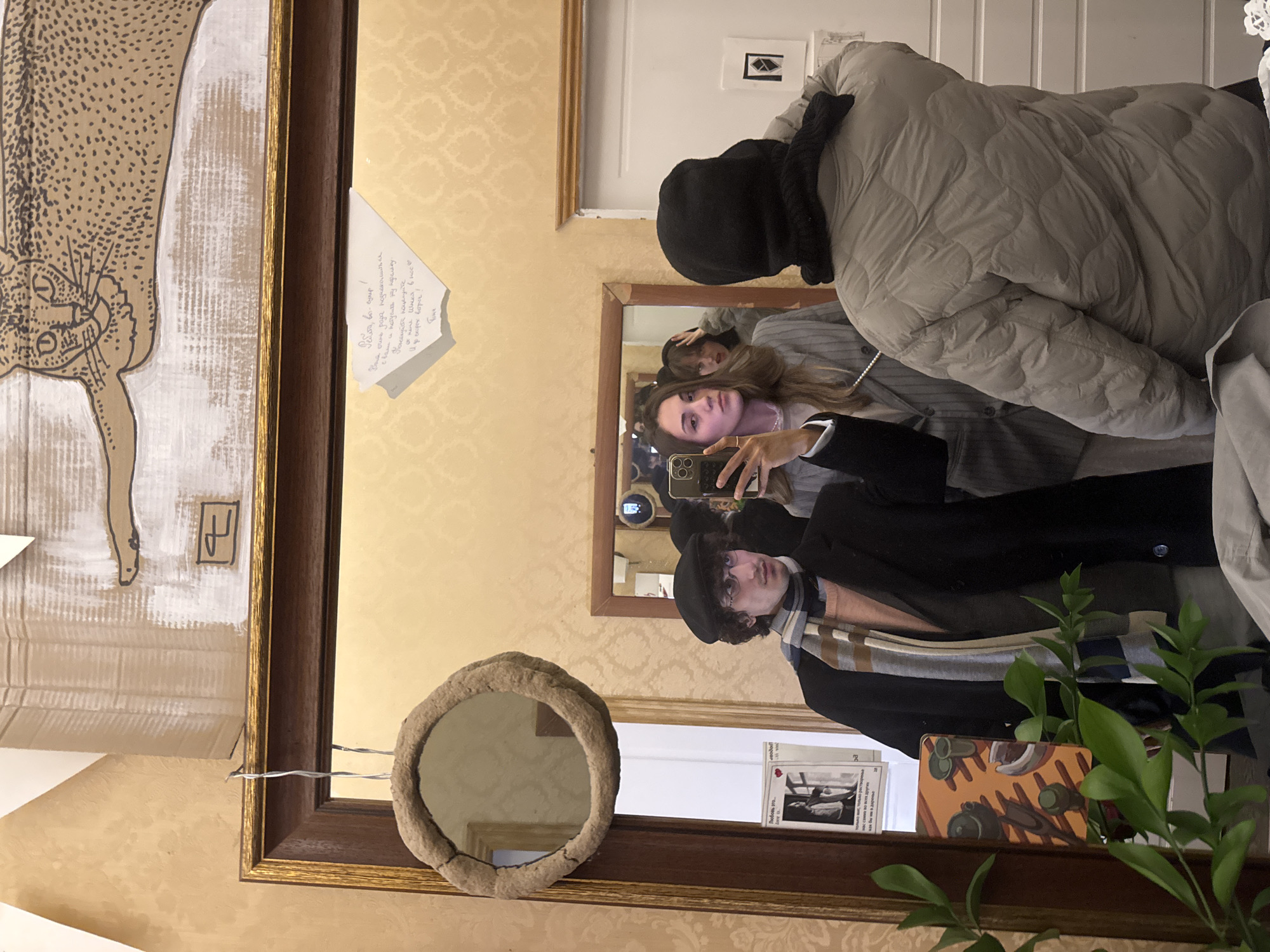 A person is taking a selfie in front of a mirror. The mirror is surrounded by artwork and decoration, including a drawing of an animal (possibly a leopard) and a smaller, round mirror. The person in the selfie is wearing a black hat and a scarf. In the reflection of the mirror, another person is visible holding a smartphone and taking the picture. The background reveals a room with patterned wallpaper and other decorative elements. A note with handwritten Russian text is attached near the artwork. There are several other objects, including decorative plants and cards, placed on the surface below the mirror. The location is likely a decorated room, possibly in a home, in Moscow, Russia, given the Russian text.