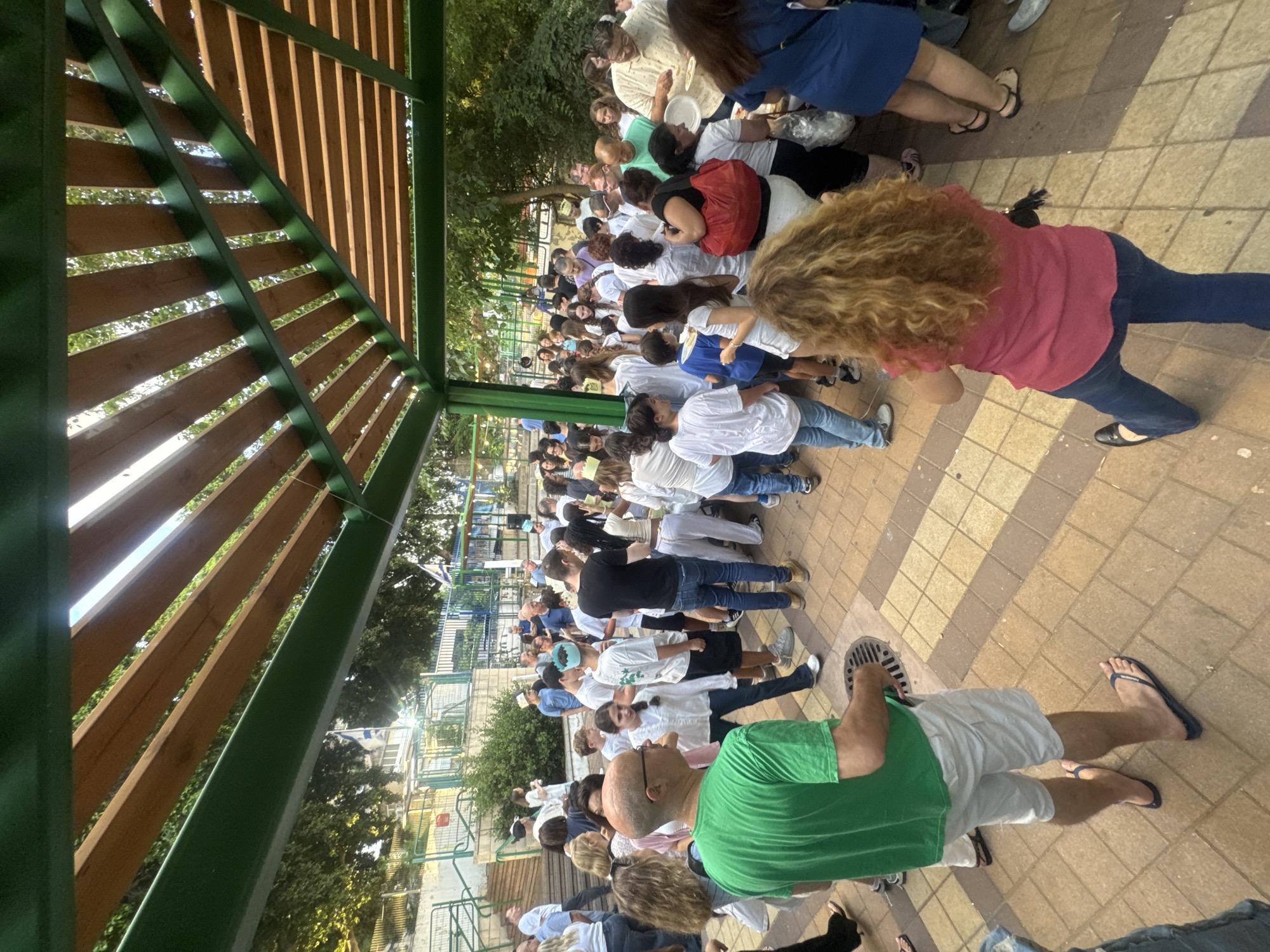 The image shows a group of people gathered outdoors, possibly for an event. The setting appears to be a paved area with some trees and buildings visible in the background. A wooden structure with green metal supports is partially visible in the foreground, casting shadows. The people are dressed in casual clothing, with many wearing white shirts. Some individuals are holding plates, suggesting a meal or a gathering. The time of day appears to be late afternoon or early evening, given the lighting and shadows. The overall mood seems relaxed and communal. No visible text is present in the image.