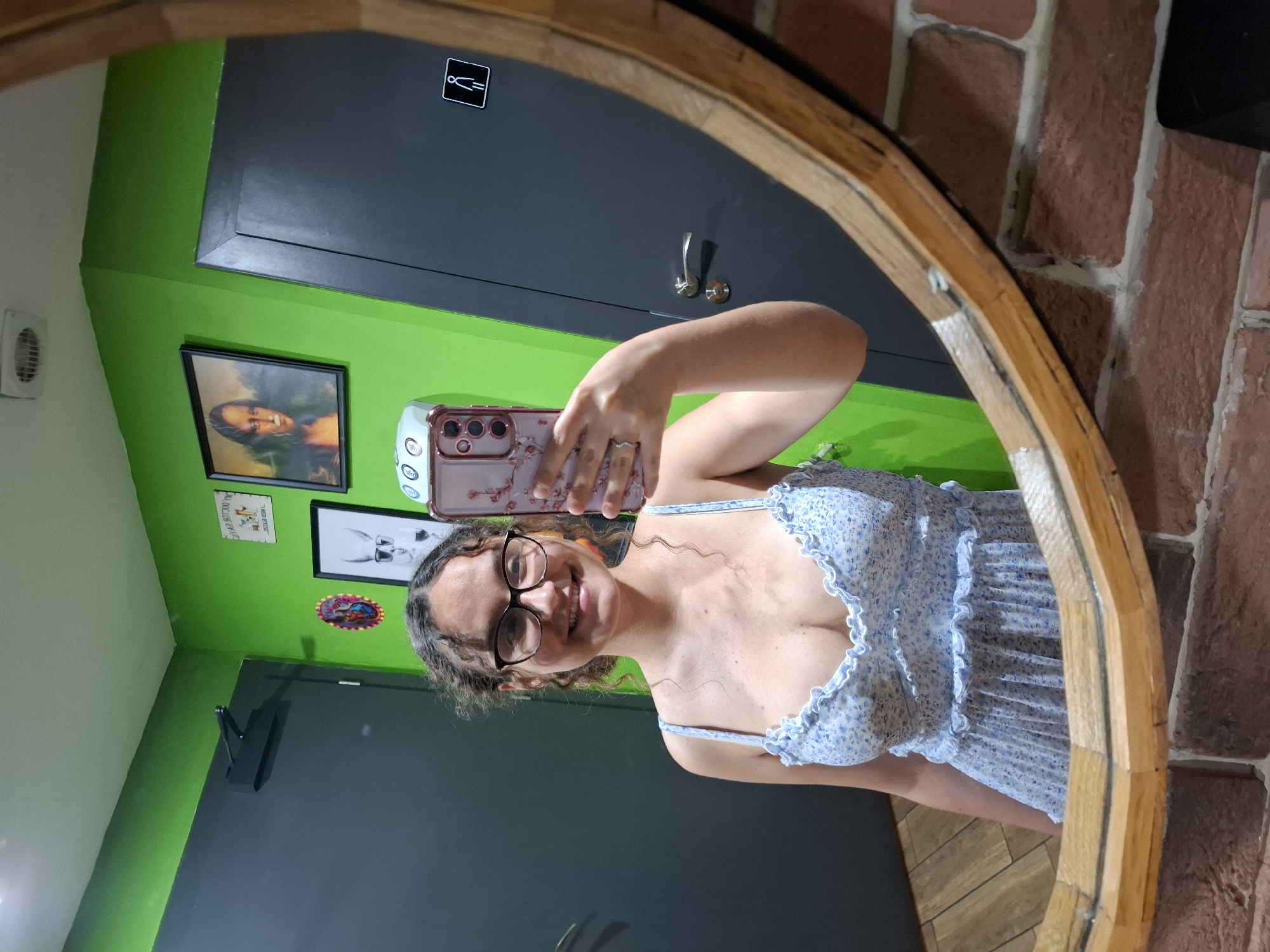 A young woman with curly brown hair and glasses takes a mirror selfie in what appears to be a public restroom. She is wearing a light blue floral print dress with ruffled straps and a ruffled hem. The restroom walls are painted a vibrant green, and there are several framed pictures on the wall, including a depiction of the Mona Lisa and a cartoon of a llama wearing sunglasses. A sign indicating accessibility is visible on a dark gray door behind her. The floor is tiled with a wood-like pattern. The mirror is framed with wood and has a rustic appearance. The woman is smiling and holding a pink smartphone with a floral case to take the photo.