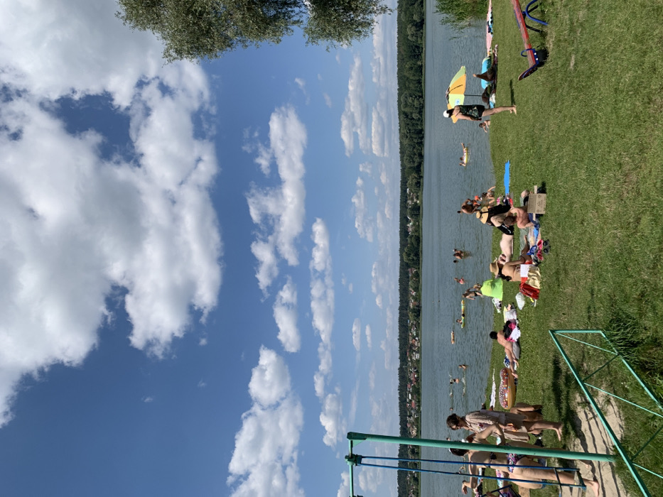 The image depicts a summer day at a lakeside beach in Mykhailivka-Rubezhivka, Ukraine, under a blue sky with scattered cumulus clouds. The central feature is a large body of calm, greenish-blue water, identifiable as Lake Mykhailivka, with a distant shoreline showing a dense line of trees and scattered residential structures. In the immediate foreground, a green grassy bank slopes towards the water. On the left, a green metal swing set is partially visible. On the right, a red and blue seesaw is present on the grass.
Approximately 25-30 individuals are observed throughout the scene. On the grassy bank, multiple individuals are sunbathing on towels and blankets; some are standing or sitting, and one person is situated under a yellow and orange beach umbrella. Personal items like bags and clothing are scattered near those sunbathing. In the lake, approximately 15-20 individuals are engaged in activities such as swimming, wading, and floating on inflatable devices, including a multi-colored donut-shaped ring. The overall scene depicts a recreational environment.